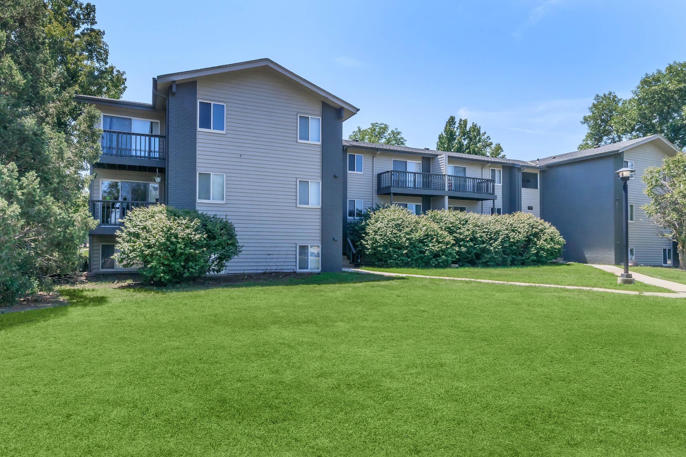 A multi-story residential building with balconies, surrounded by lush green grass and shrubs under a clear blue sky. The structure features a mix of siding colors, with trees in the background, creating a pleasant, inviting atmosphere.