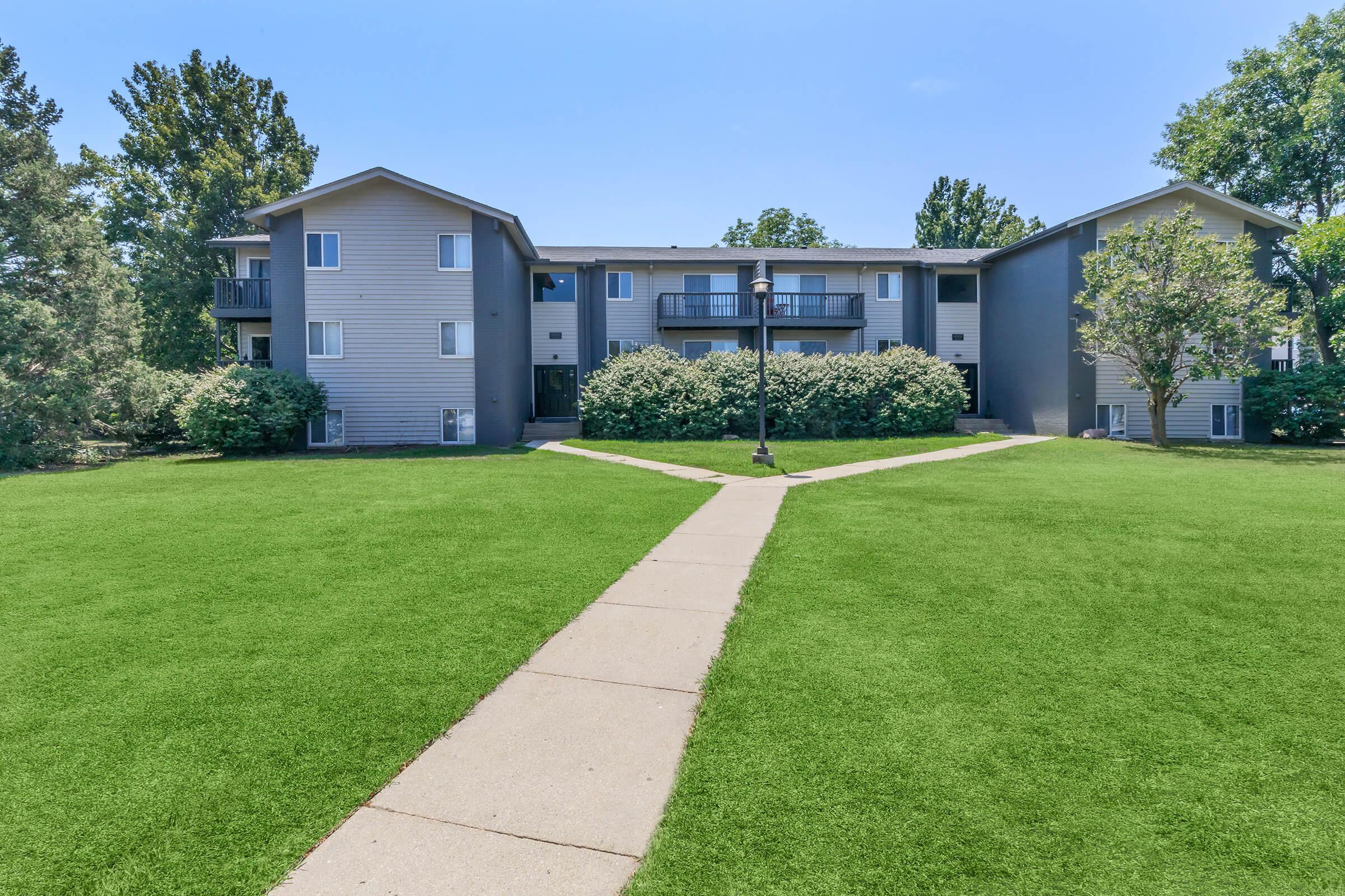 A well-maintained apartment complex with three buildings surrounded by green grass and shrubs. A paved pathway leads through the lawn, connecting the buildings. Trees are visible in the background under a clear blue sky. Ideal for showcasing residential living spaces.