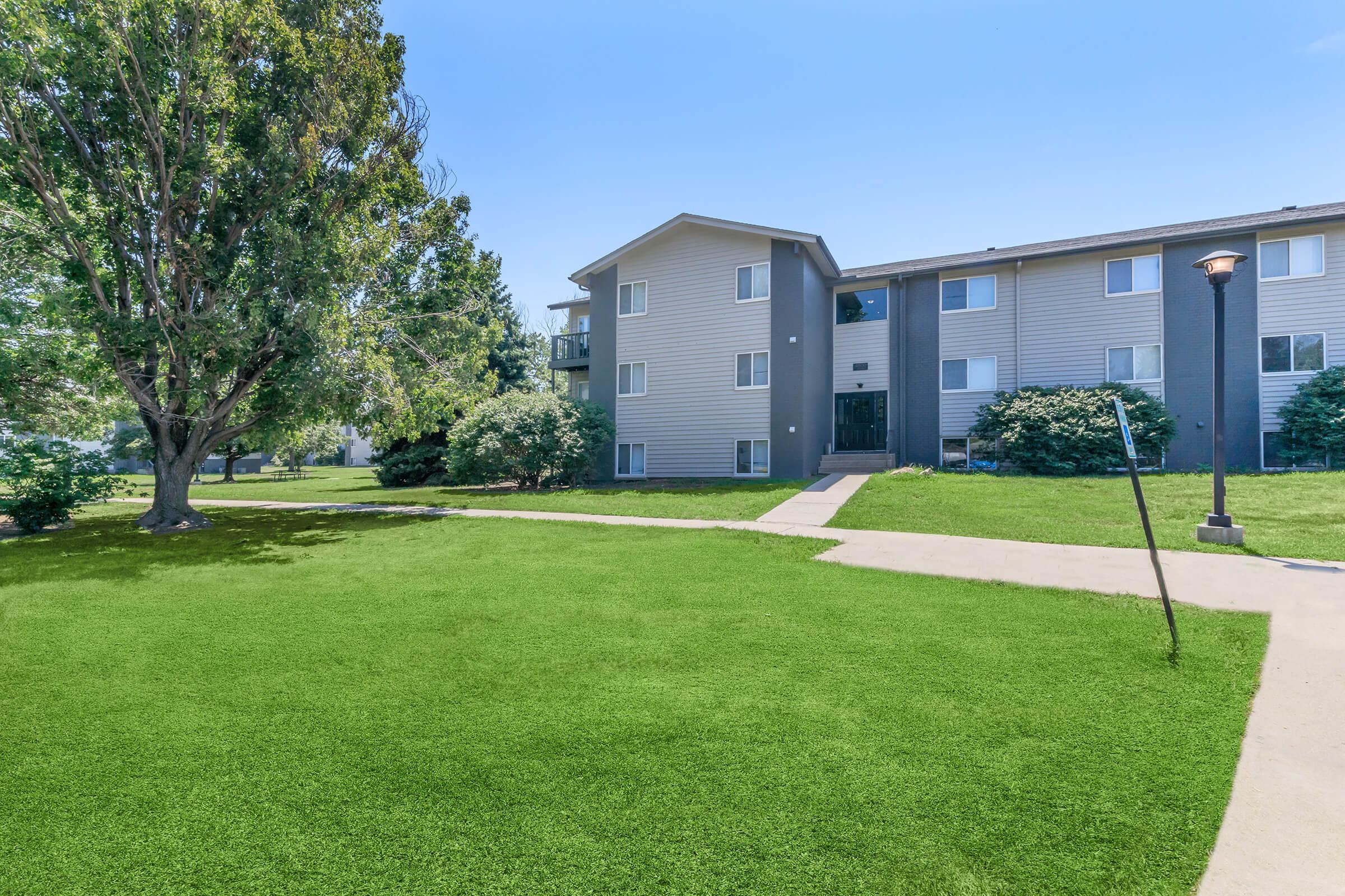 Apartment building with multiple floors, surrounded by green grass and trees. A pathway leads to the entrance, and a lamppost stands nearby. The sky is clear and blue, contributing to a bright, inviting atmosphere.
