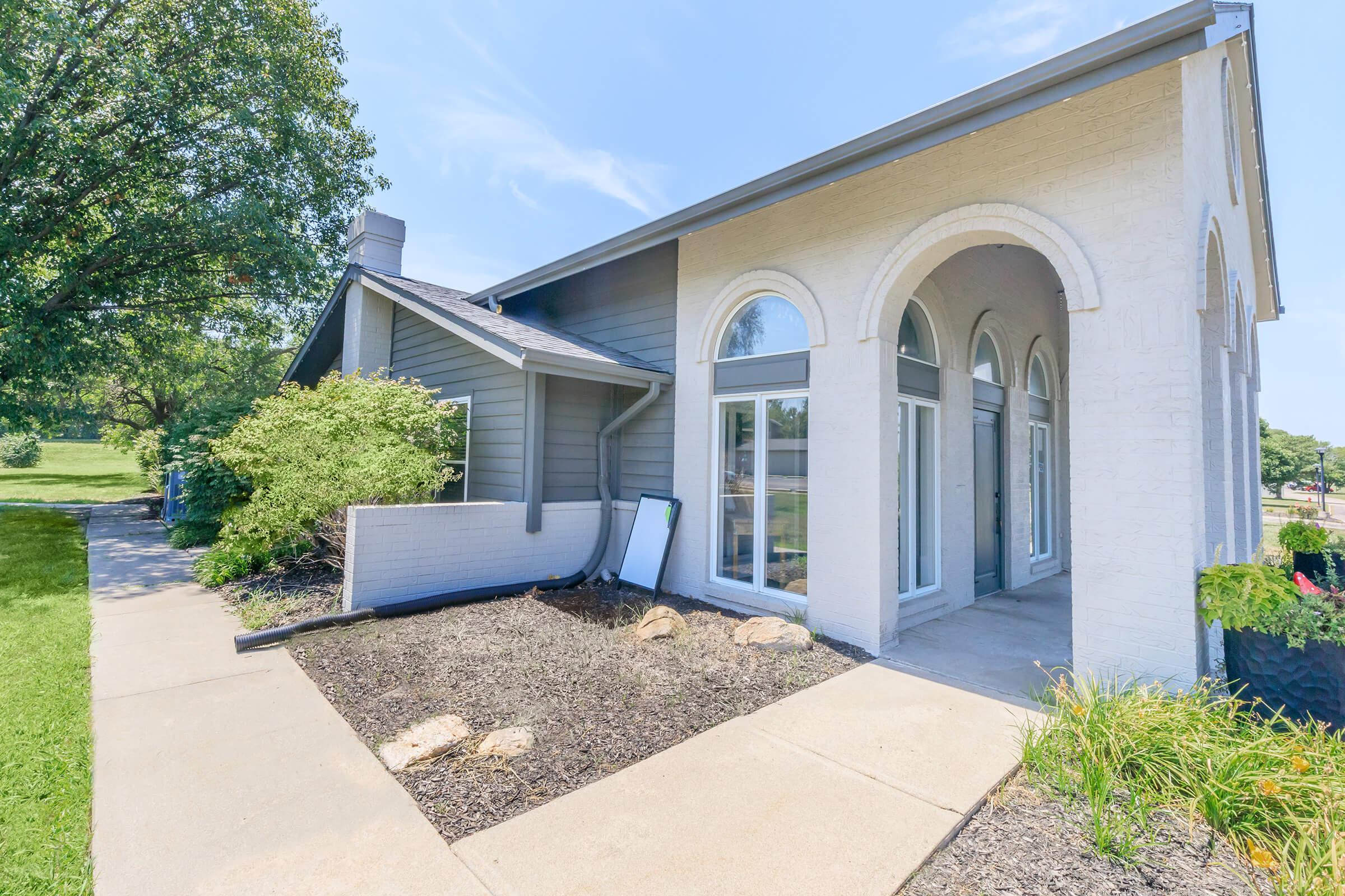 A modern building with a combination of gray and white exterior, featuring large arched windows and a landscaped front area. The path is lined with rocks and small plants, and there are trees in the background under a clear blue sky.
