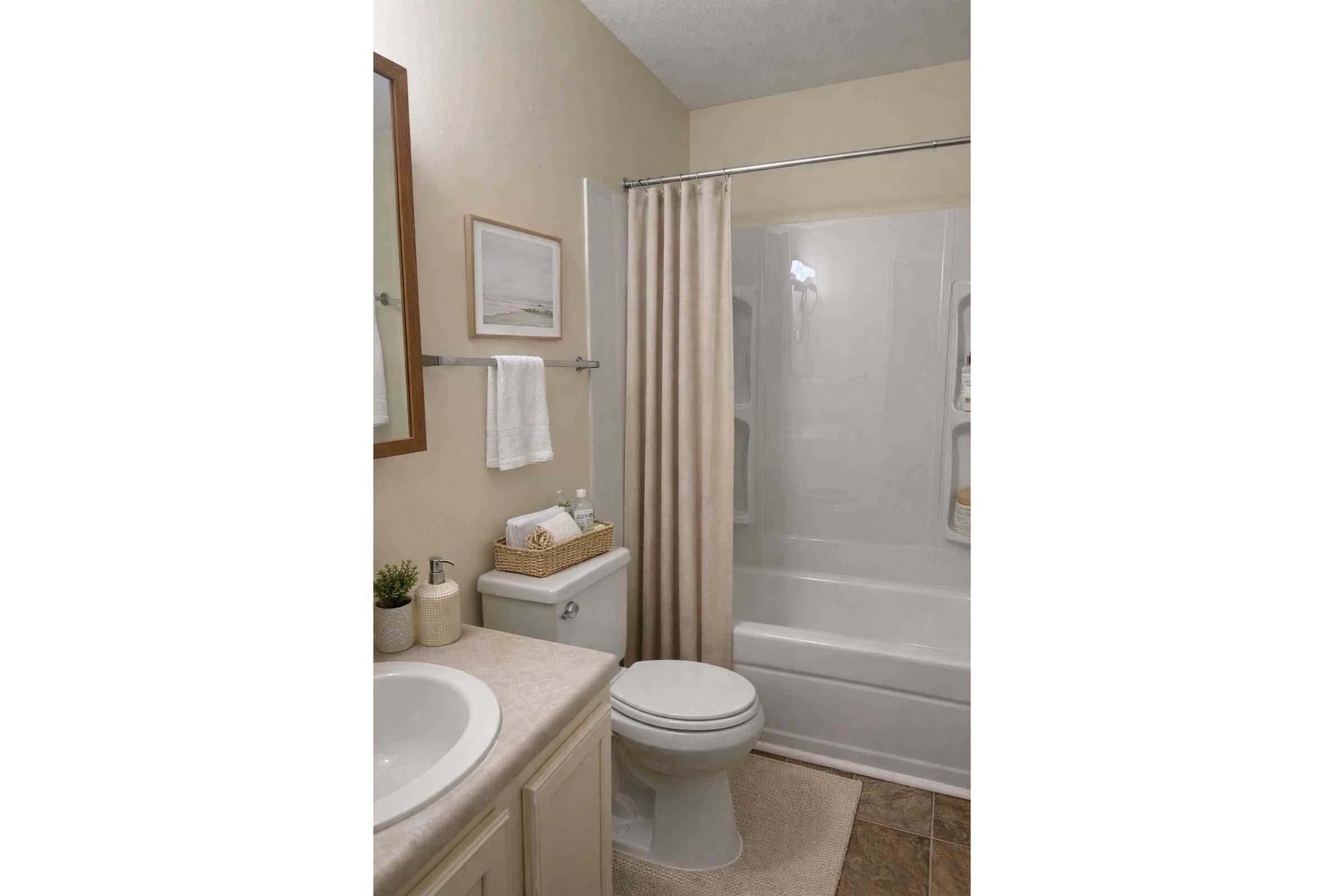A well-organized bathroom featuring a white bathtub with a glass shower curtain, a toilet, and a sink with a countertop. Decor includes a small potted plant, framed artwork, towels, and toiletries neatly arranged on a shelf. Neutral color palette enhances the calm ambiance.
