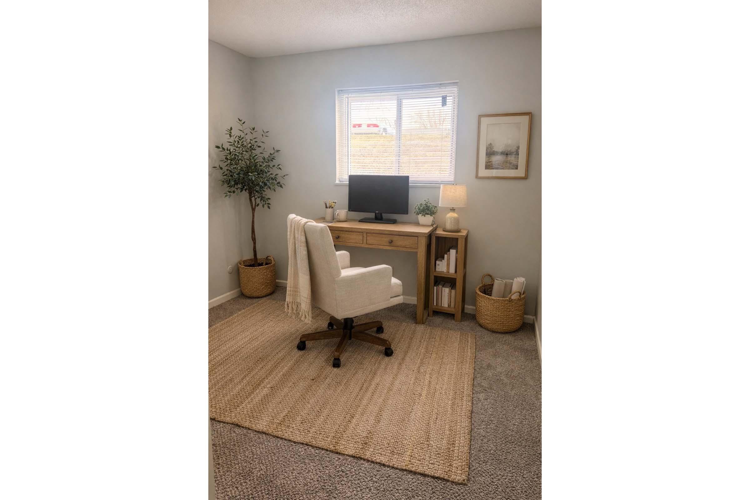 A cozy home office featuring a wooden desk with a computer monitor, a comfortable white chair, and a small lamp. Decorated with a framed picture on the wall, a potted plant in the corner, and a woven basket holding supplies. The space is well-lit with natural light from a window, and has a neutral, inviting color scheme.