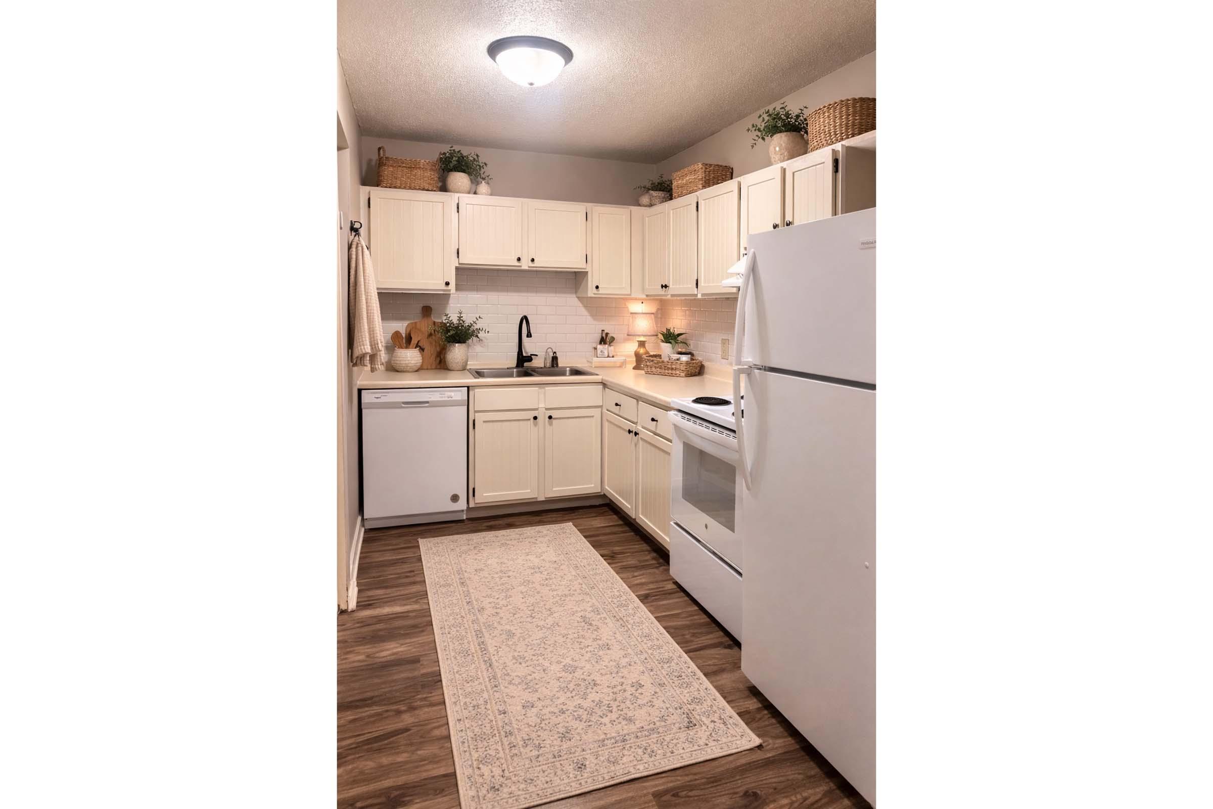 A modern kitchen showcasing white cabinetry, a countertop with a sink, and essential appliances including a refrigerator and an oven. The space is decorated with green plants and decorative baskets, complemented by a light decorative rug on the floor, creating a warm and inviting atmosphere.