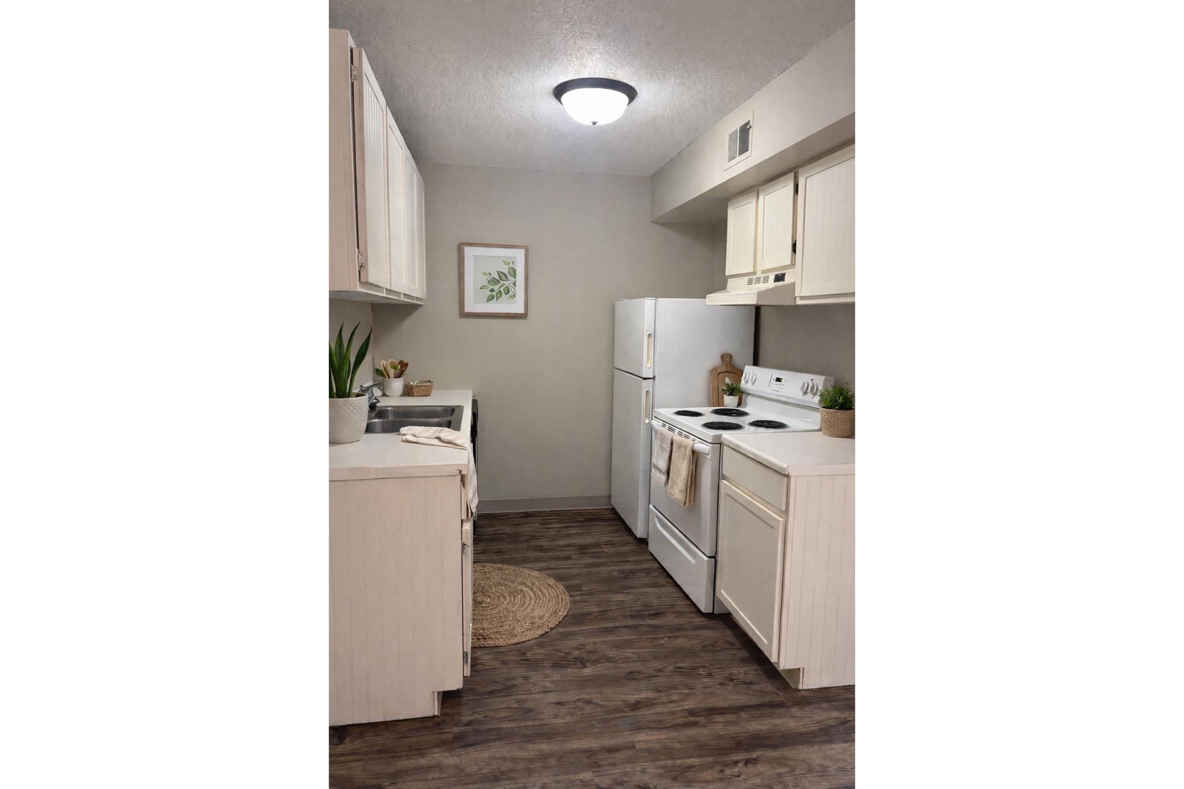 Modern kitchen with light-colored cabinets, white appliances, and wooden flooring. There is a small round rug on the floor, a plant on the counter, and framed artwork on the wall. The overall design is bright and inviting, showcasing a functional and stylish cooking space.