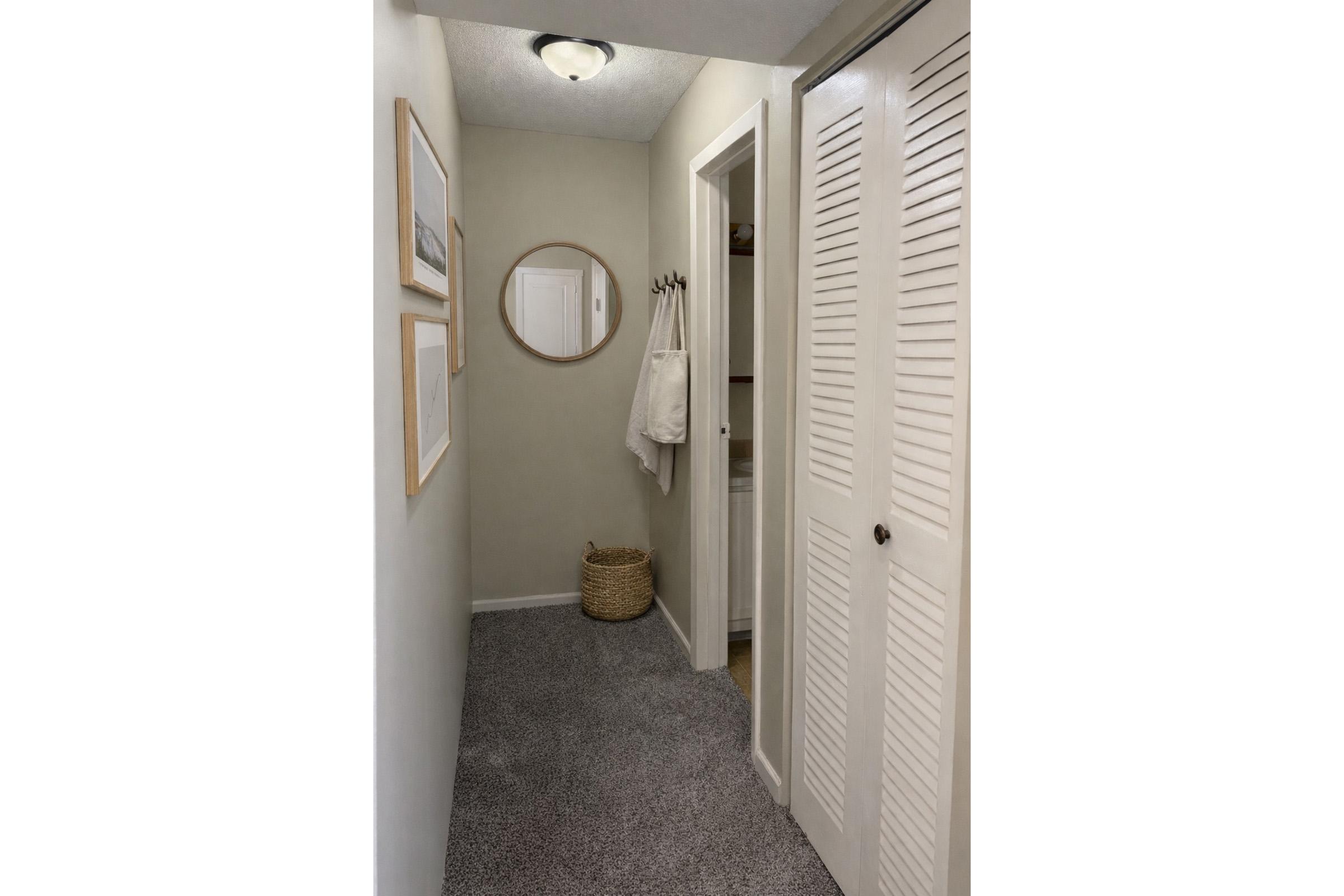 A narrow hallway featuring neutral wall colors, a round mirror, two framed artworks, a closet with louvered doors, and a beige woven basket on the floor. A towel hangs on a hook near the closet, and the space is illuminated by a ceiling light. The carpet is light gray.