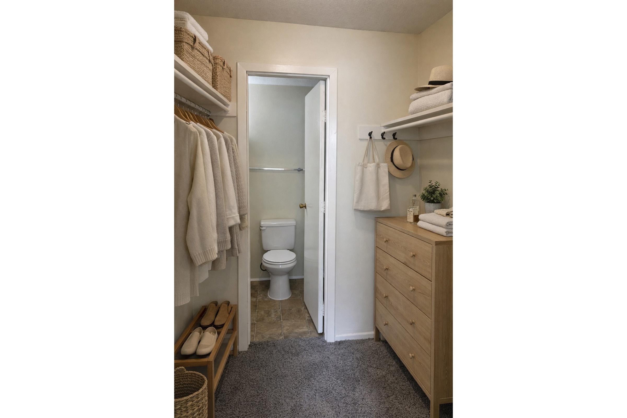 A neatly organized bathroom featuring a small closet space with hanging white towels, a toilet in the background, and a wooden storage unit with decorative items. Soft lighting highlights the clean, minimalistic decor, contributing to an inviting ambiance.