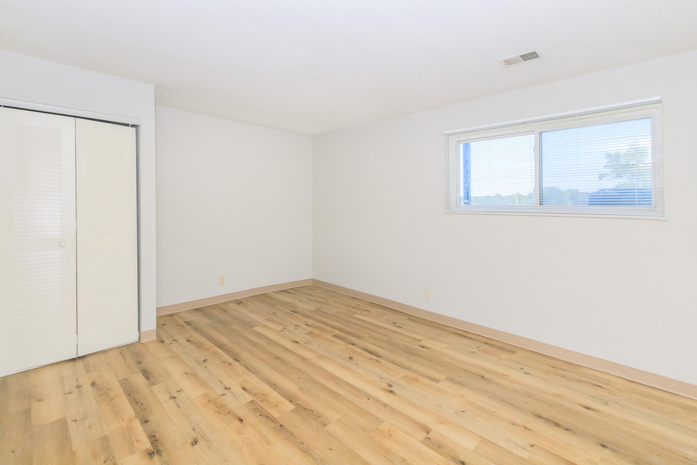 A bright, empty room with light-colored walls and a large window. The floor is wooden with a light finish. There's a closet with sliding doors on one side of the room. The space is well-lit, suggesting a clean and inviting atmosphere, perfect for customization or personal decoration.