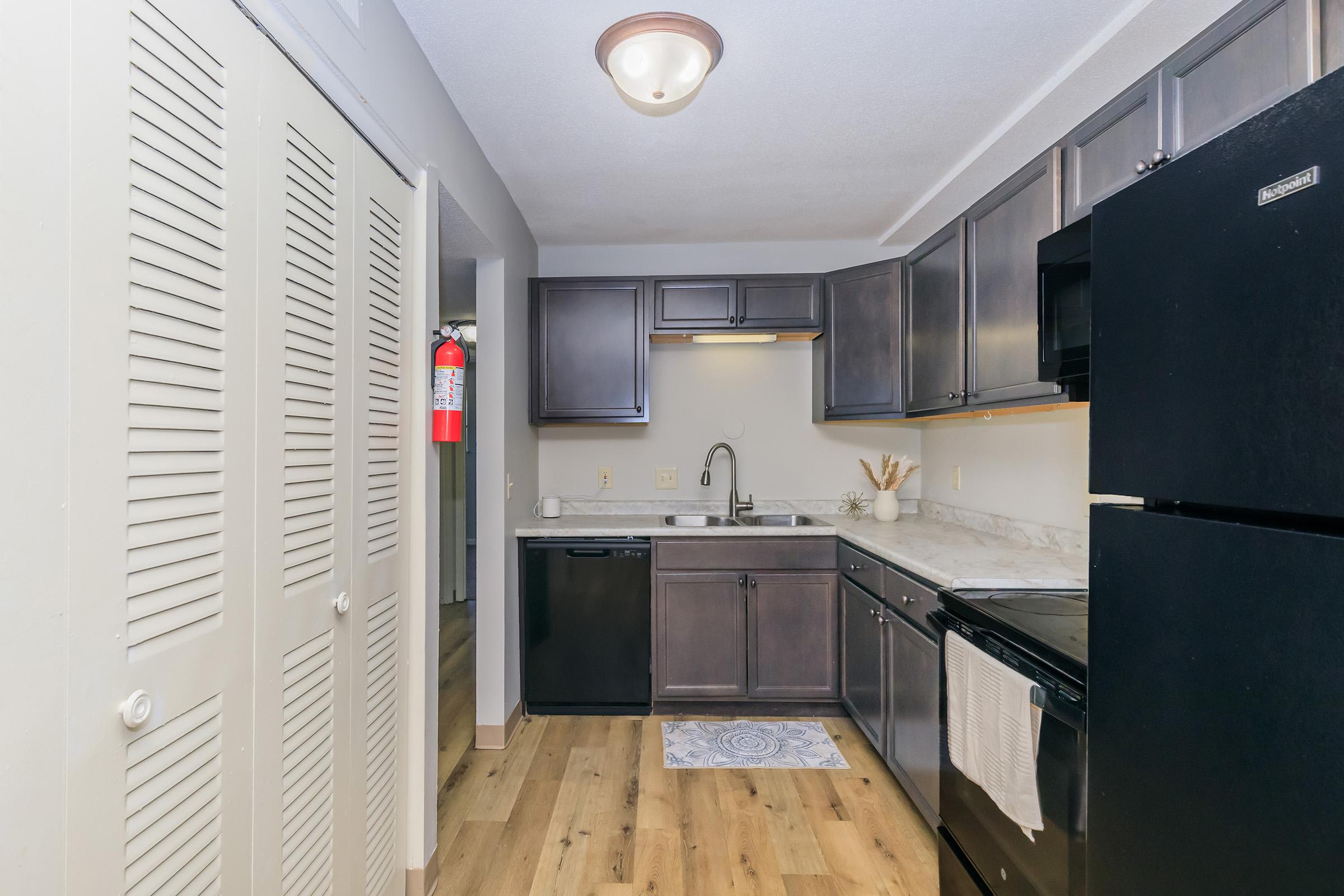 A modern kitchen featuring dark wood cabinets, a black refrigerator, and a black stove. The countertop is light-colored with a sink and dish mat. There are closed louvered doors on one side, and a fire extinguisher is visible on the wall. The floor is wood laminate, creating a warm and inviting atmosphere.