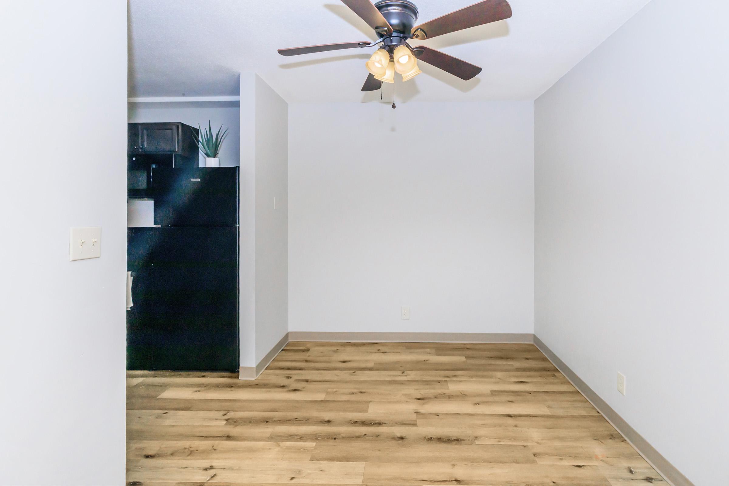 Empty room with light-colored walls and wooden flooring. A ceiling fan with light fixtures is visible, and at the back, there is a black refrigerator. The space is well-lit, creating a clean and open atmosphere.