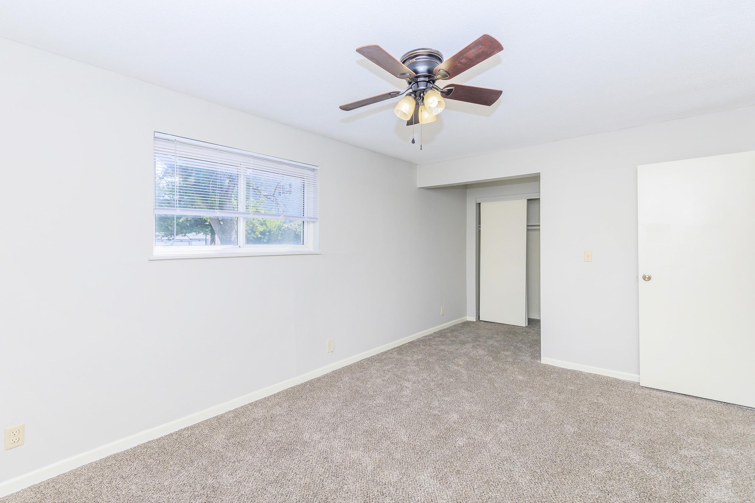 A spacious room with light-colored walls and carpeted flooring. A ceiling fan with a light fixture hangs from the ceiling. There is a window allowing natural light, and a doorway leading to a closet or additional space. The room is empty, creating a fresh and airy atmosphere.