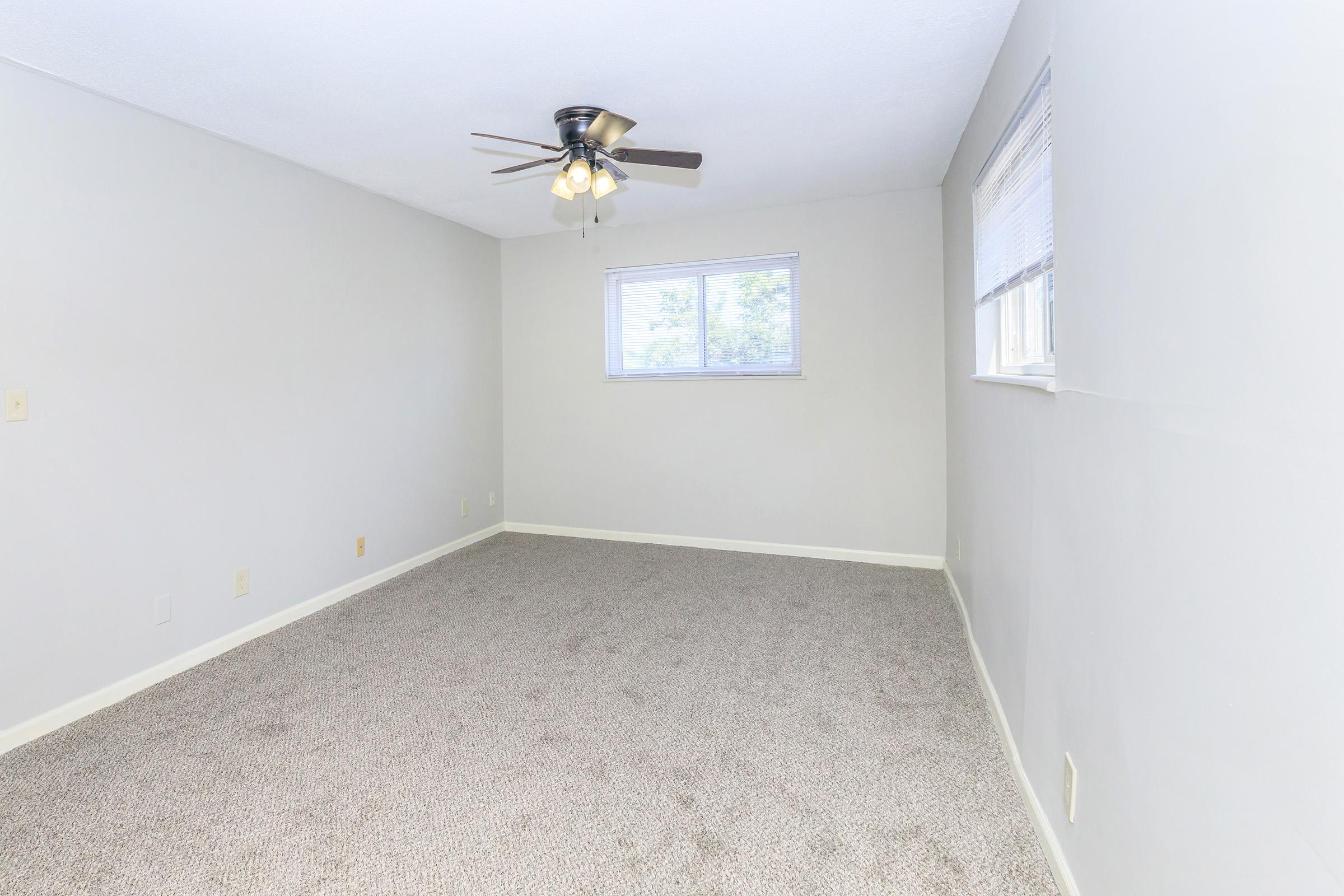 A bright, empty room with light gray walls and plush beige carpet. A ceiling fan with four light bulbs hangs from the ceiling. There is a window on one wall allowing natural light to enter. The space is clean and uncluttered, creating an inviting atmosphere.