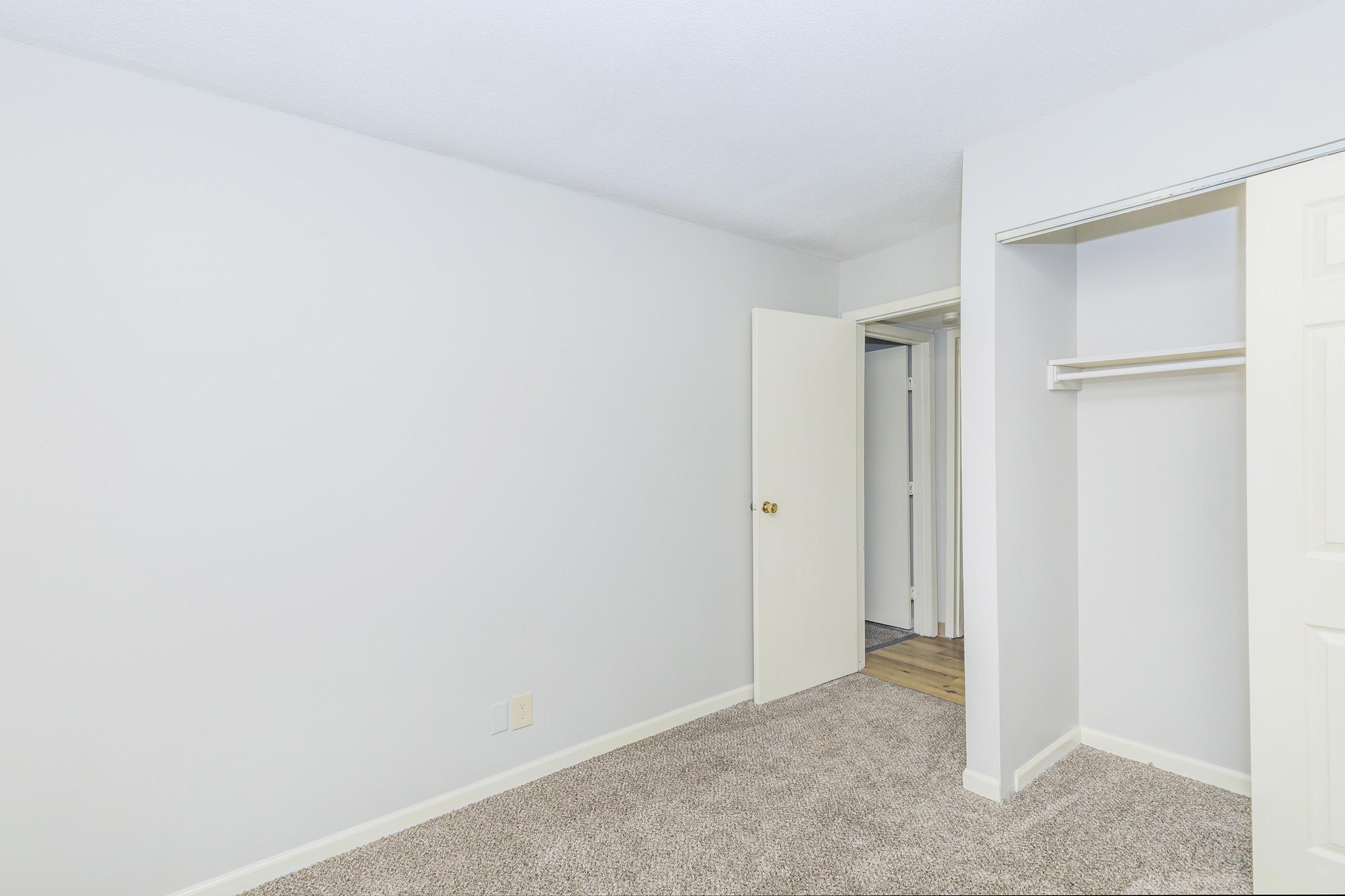 A clean, empty bedroom with light gray walls and plush carpet. A white sliding closet door is partially open, revealing shelving. An open door leads to a small hallway, indicating access to another area. The overall atmosphere is bright and spacious, suitable for personalization.