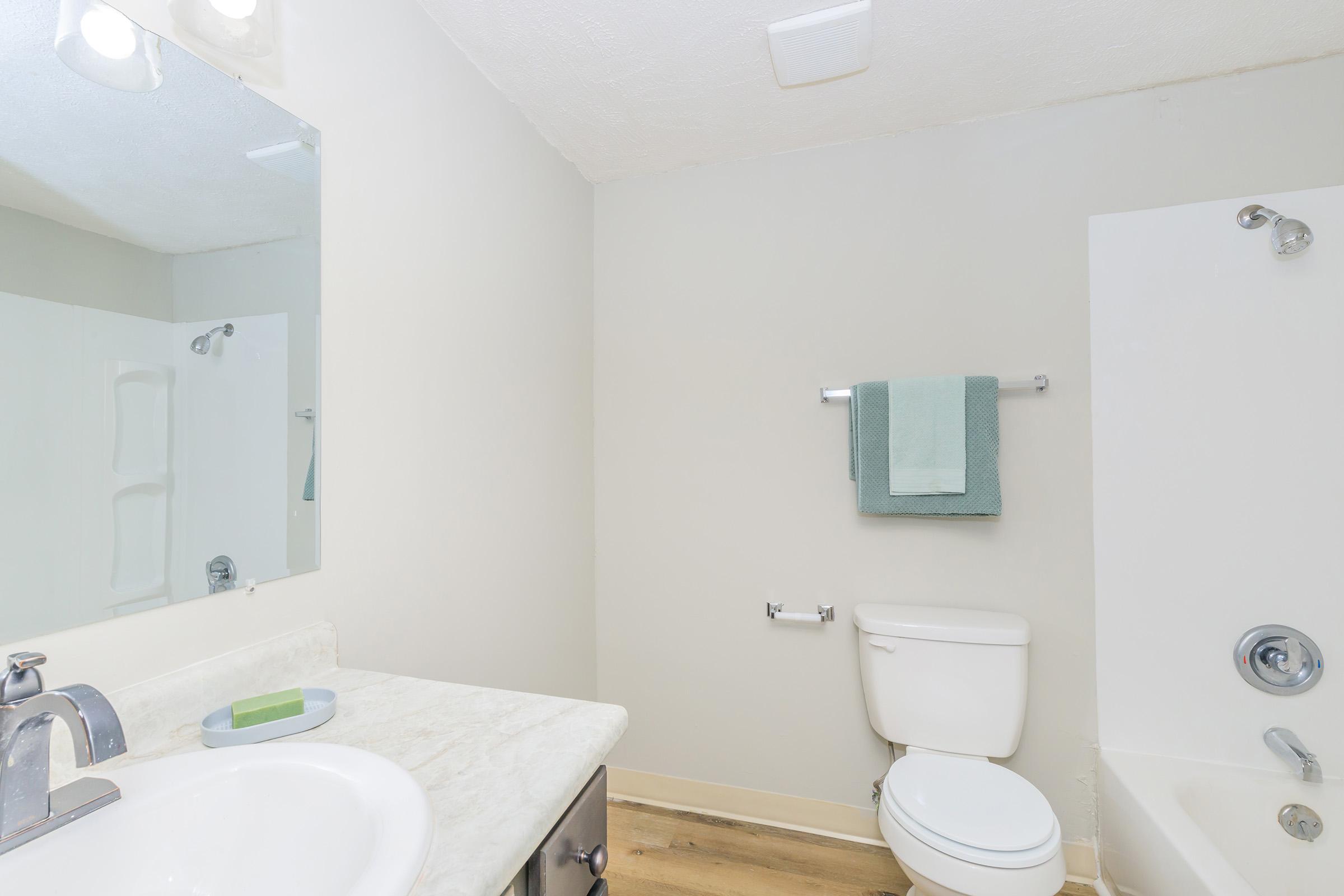 A clean and modern bathroom featuring a white sink with a light-colored countertop, a toilet, and a bathtub with a shower. The walls are painted light gray, and there is a mirror above the sink. A green towel and washcloth hang on a bar next to the sink, and a small green soap dish is on the countertop.