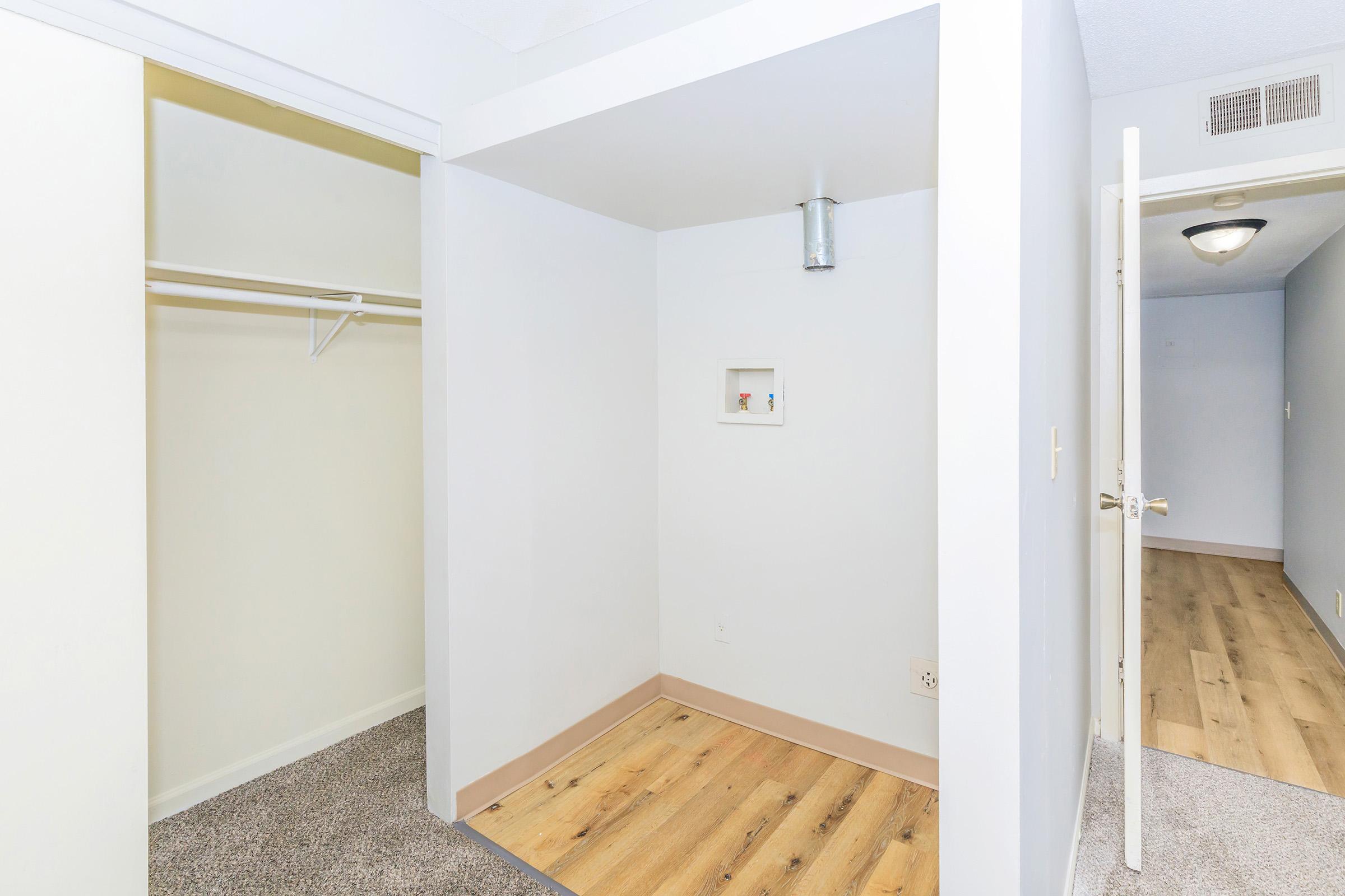 A small, empty room with light-colored walls and wooden flooring. To the left, there is an open closet with a hanging rod. On the right, a doorway leads to another area, and there is a ceiling light visible. The space appears clean and uncluttered, suitable for storage or as a small office.