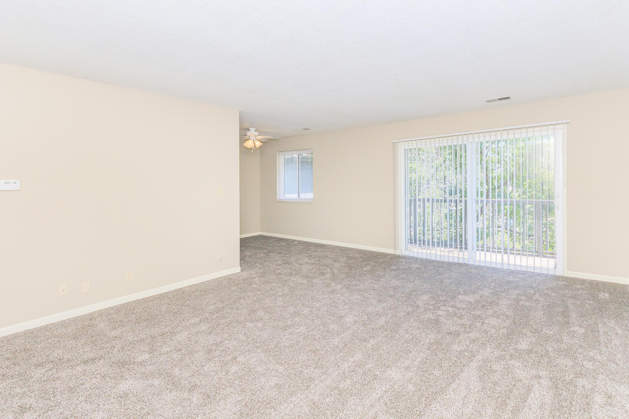 Spacious empty living room featuring light beige walls, a ceiling fan, and soft carpet. Large sliding glass door leads to a balcony with greenery visible outside. Natural light fills the room, creating a bright and airy atmosphere.