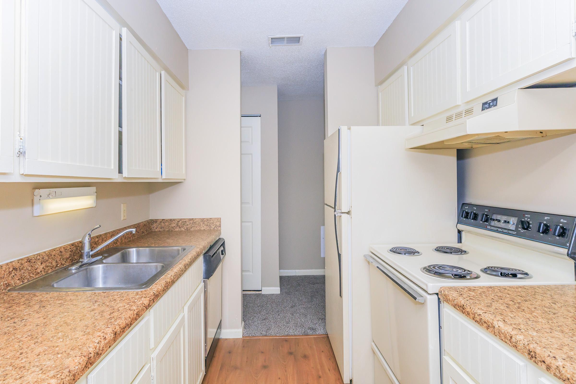 A bright kitchen featuring white cabinetry, a large countertop made of light-colored stone, an oven and stove, and a refrigerator. There is a stainless steel sink with a single faucet. The kitchen leads to a hallway with a door visible at the end. The walls are painted in a neutral tone, and the floor has a laminate finish.