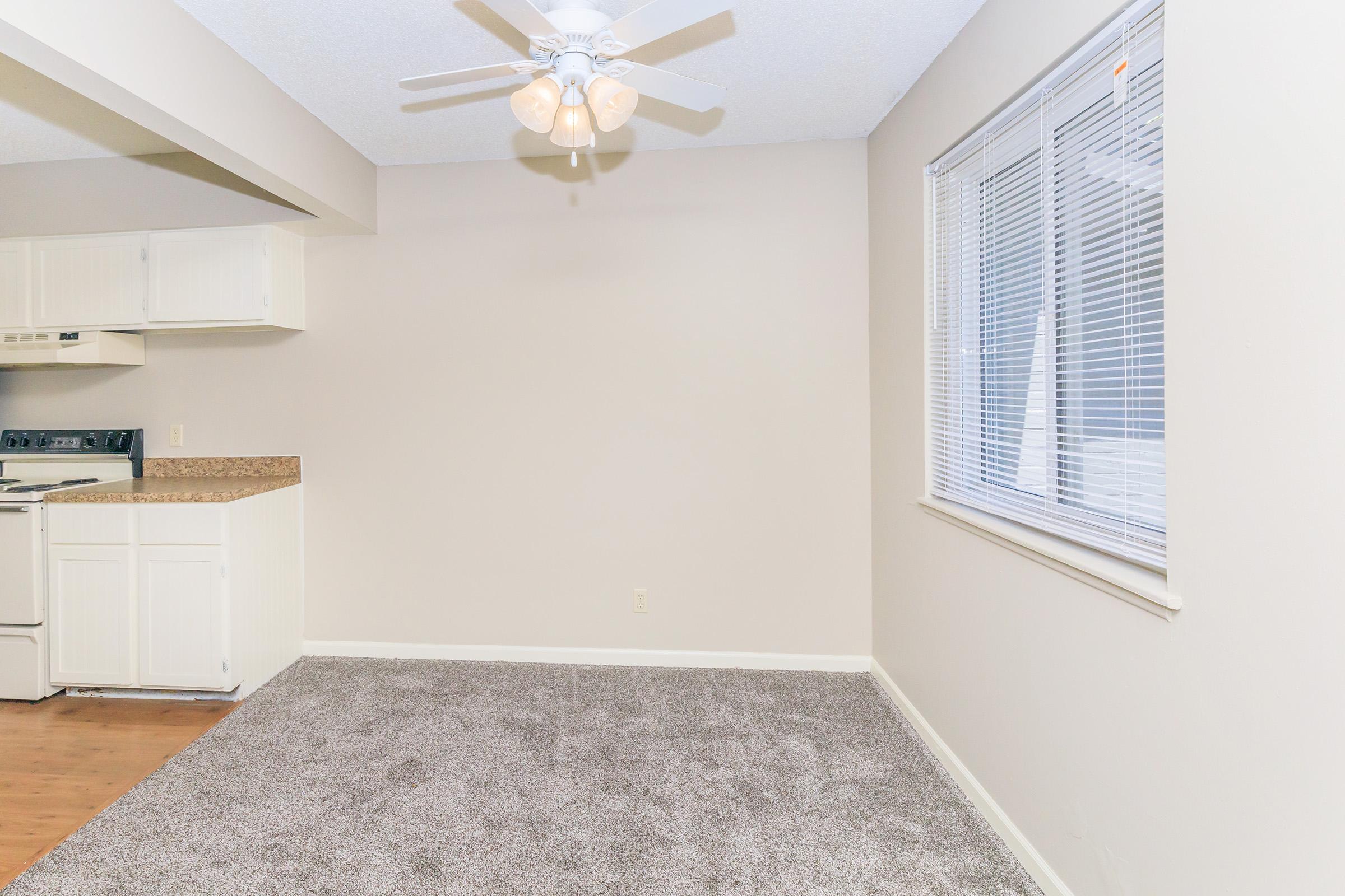 A spacious room with light beige walls and a ceiling fan. The floor is carpeted in a light gray, and there is a window with blinds allowing natural light. A partially visible kitchen area features white cabinetry and a stove. The overall ambiance is clean and inviting.