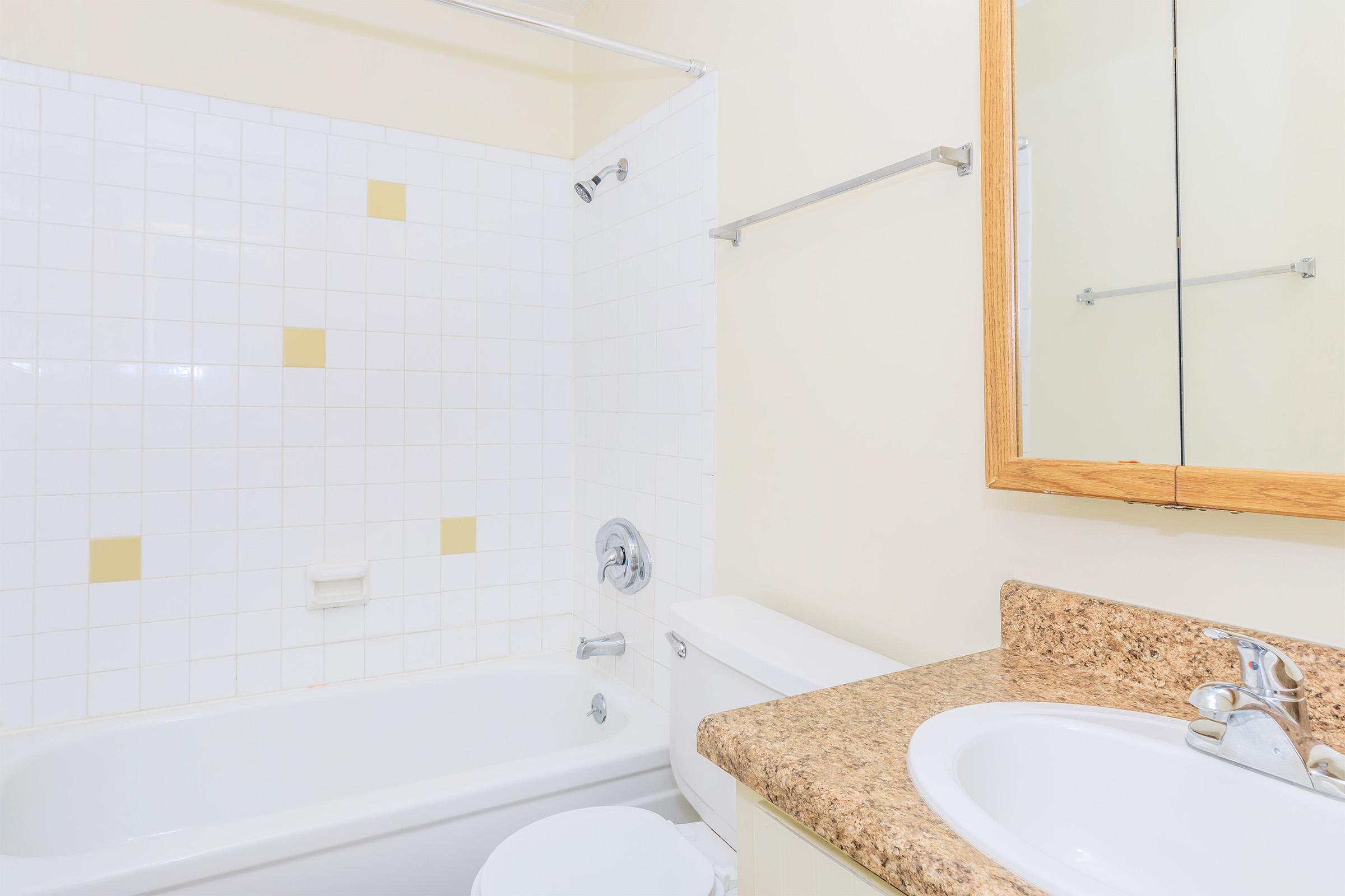 A clean, well-lit bathroom featuring a bathtub with a shower, white tiled walls with yellow accents, a beige countertop with a sink, and a mirror above. The space is tidy and minimalistic, emphasizing a neutral color palette.