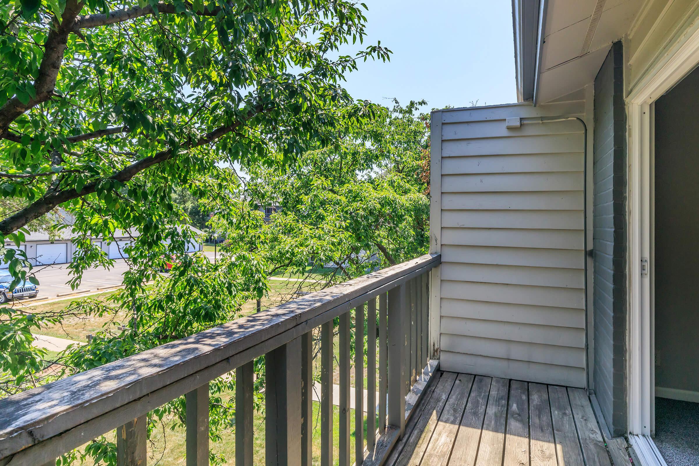 View from a balcony looking out onto greenery and a pathway. The wooden railing frames the image, with leaves from nearby trees creating a natural setting. A corner of a building is visible, suggesting a residential area. The scene is bright and inviting, with clear blue skies above.