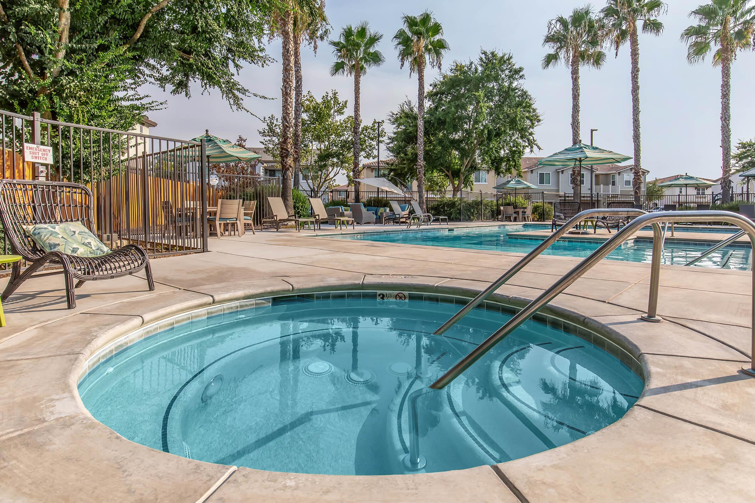 Apartments for Rent in Fresno County, CA - Parador Townhomes - Soothing Spa and Simmering Swimming Pool With Lounge Tanning Chairs, Umbrellas, Tables, and Chairs Surrounded by Palm Trees
