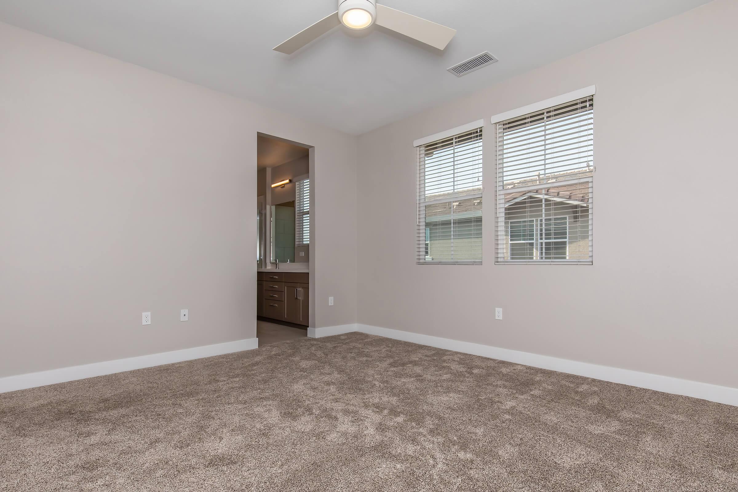 Fresno County, CA, Apartments - Parador Townhomes - Empty Bedroom with Plush Light Brown Carpet, A Ceiling Fan, Two Large Windows With Blinds, With Direct Acess To The Bathroom
