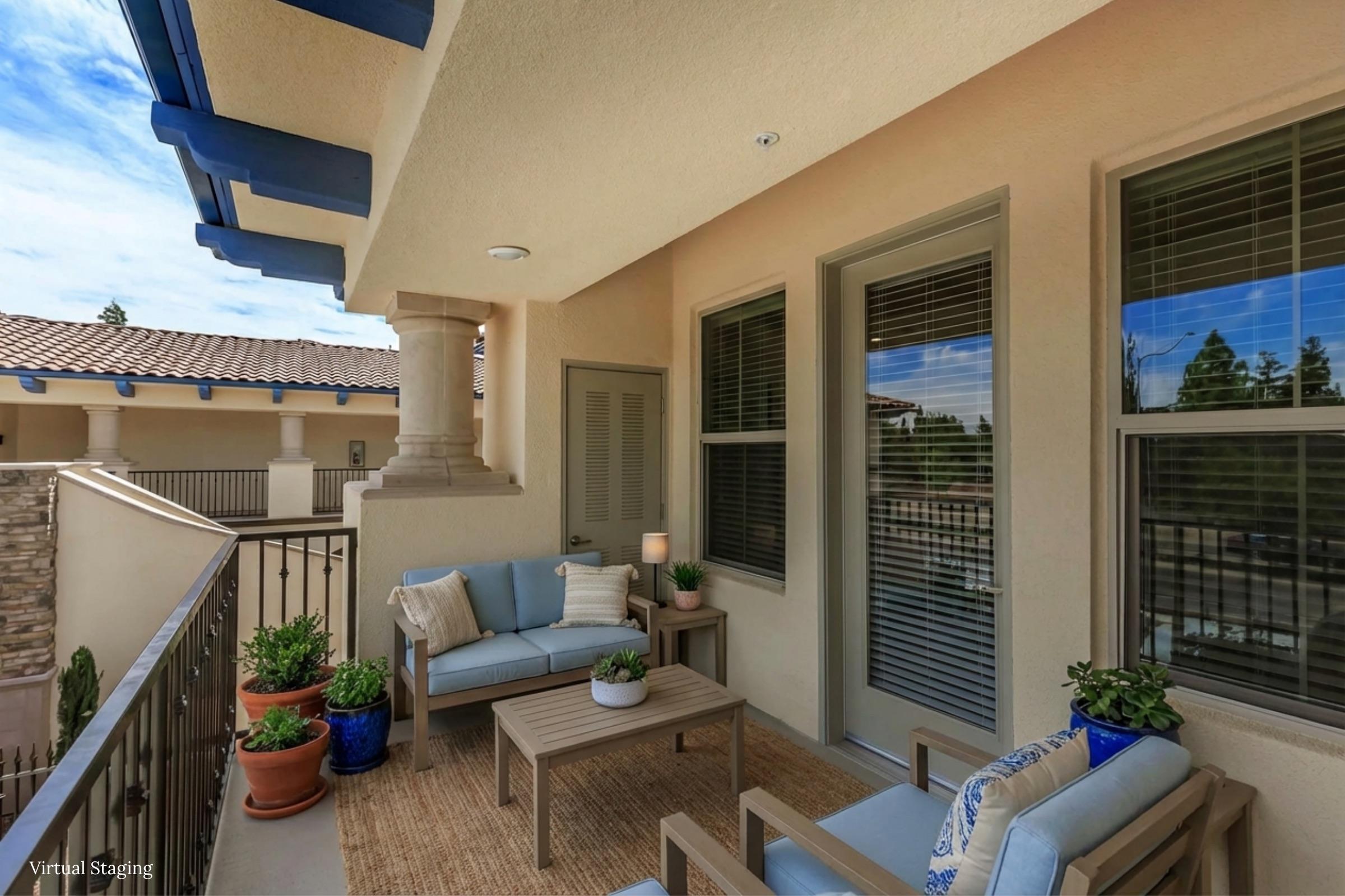 A cozy patio area featuring a small table and two comfortable chairs, surrounded by potted plants. Large windows with blinds allow natural light. The space is adorned with neutral colors and modern decor, creating a welcoming atmosphere for relaxation.