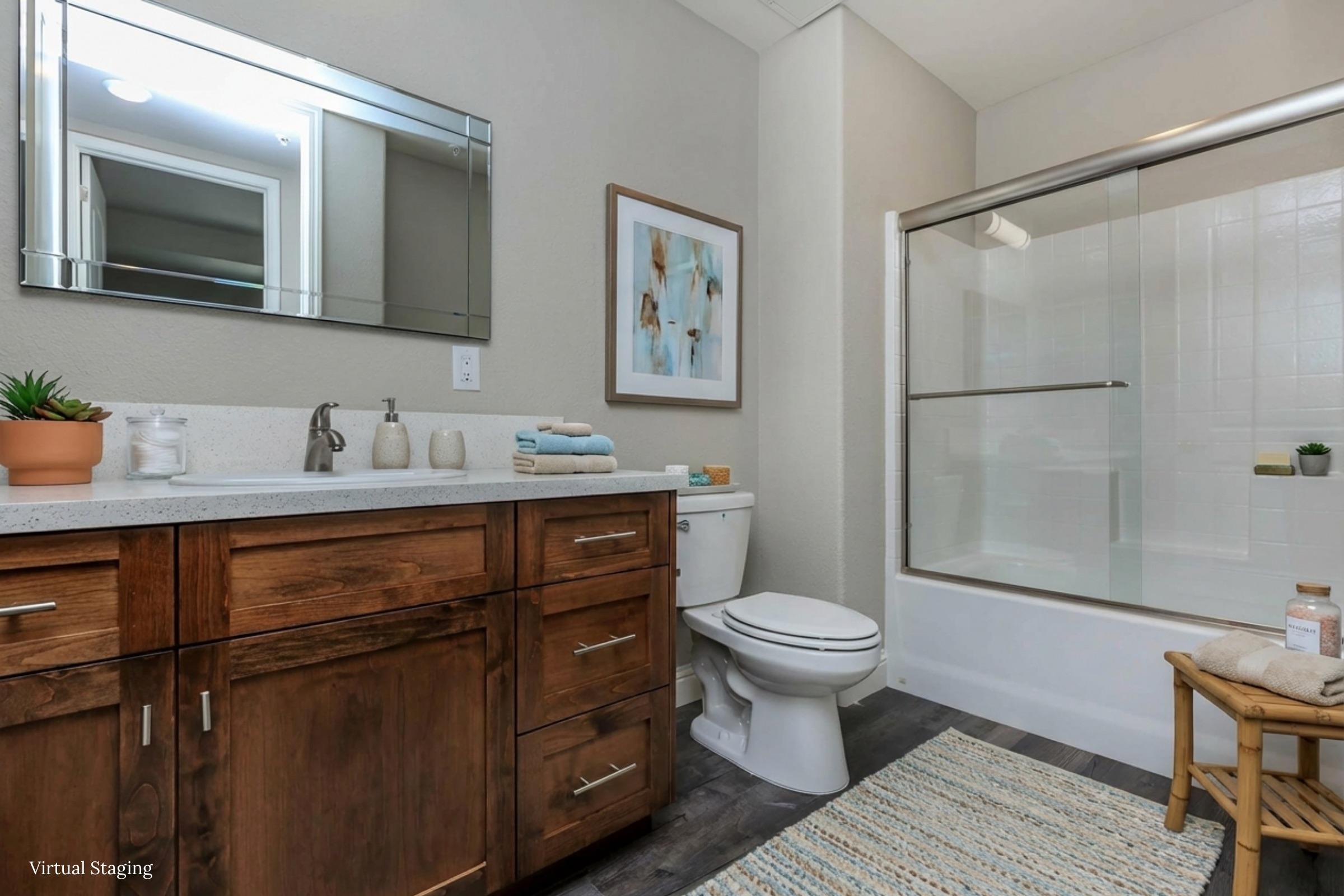 A modern bathroom featuring a wooden vanity with a sink, a large mirror, a toilet, and a glass-enclosed shower. Neutral wall colors and decor add a contemporary feel, complemented by a small plant and neatly stacked towels. A woven rug enhances the overall aesthetic.