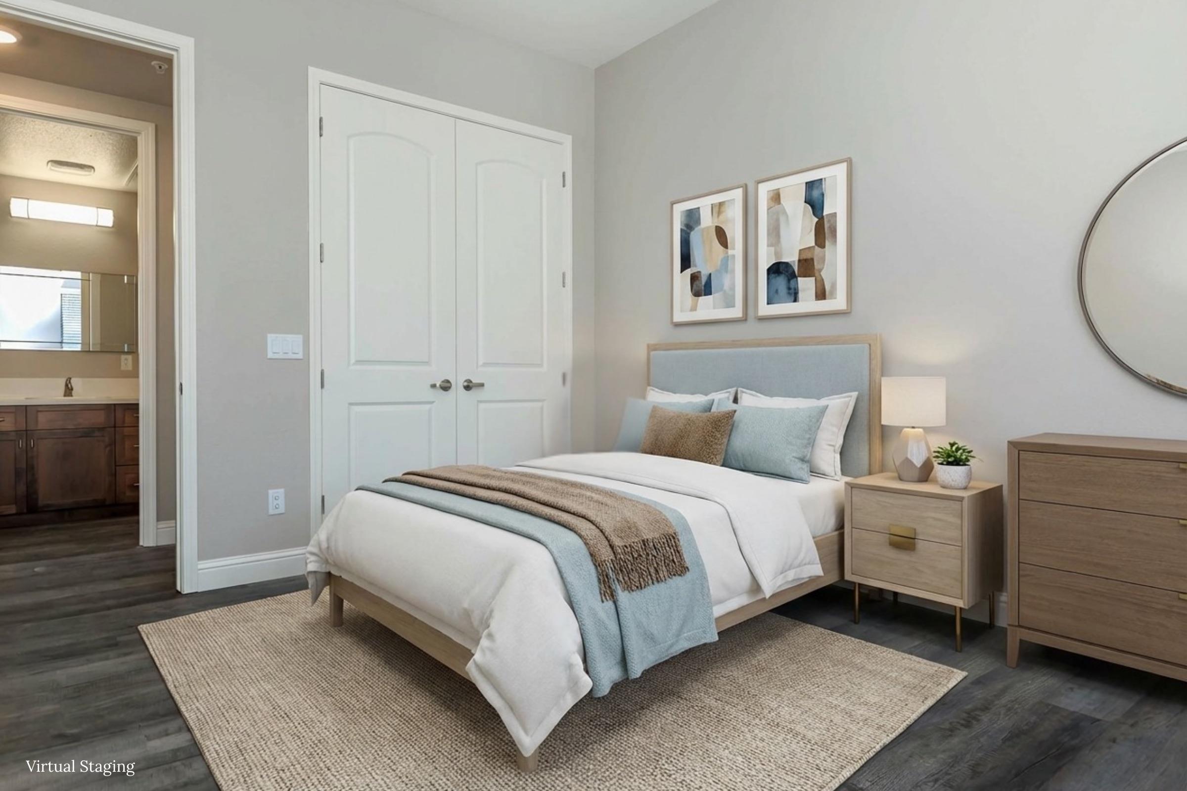 A cozy, modern bedroom featuring a light-colored bed with a blue and white bedding set, a wooden nightstand with a lamp, and artwork on the walls. A round mirror is positioned above the nightstand, and a natural fiber rug covers the floor. The room is well-lit with neutral walls and an open doorway leading to a bathroom.