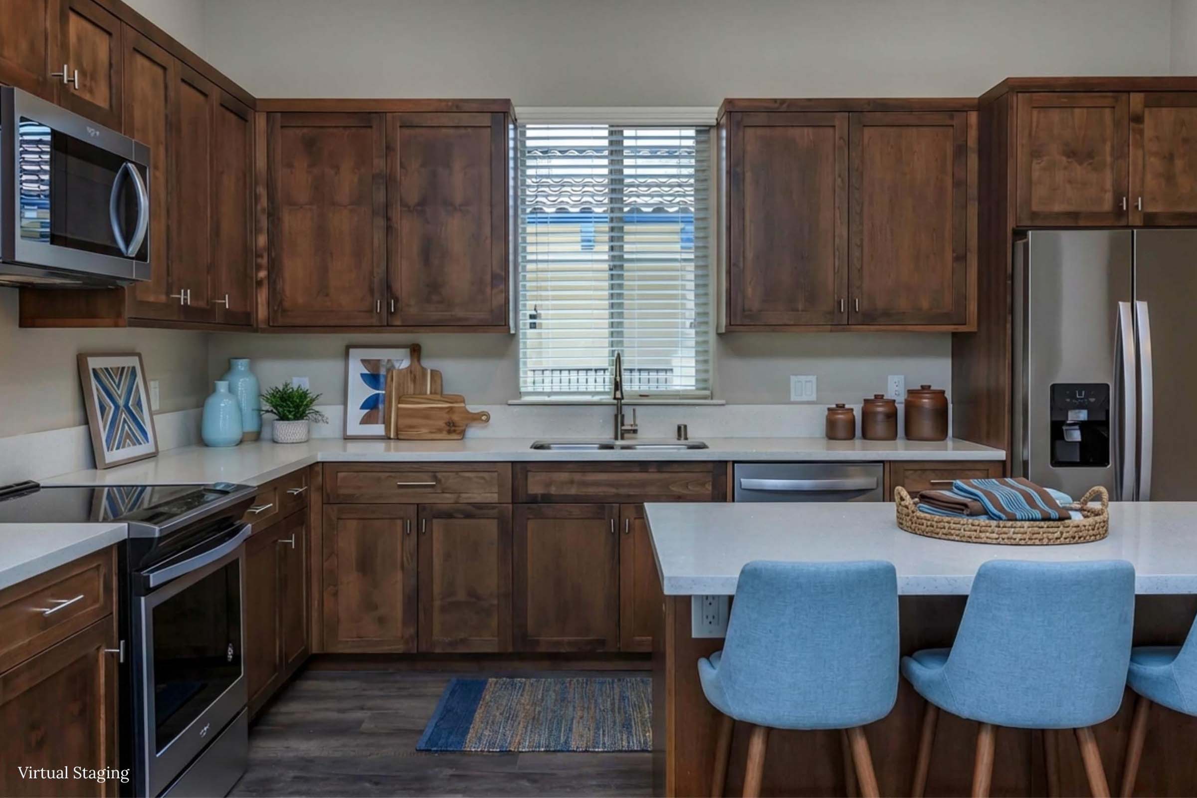 Modern kitchen featuring wooden cabinets, stainless steel appliances, and a white countertop. A window brings in natural light, and a small blue rug adds color. The space includes two light blue bar stools at the counter and decorative items on the shelves, creating a cozy and inviting atmosphere.