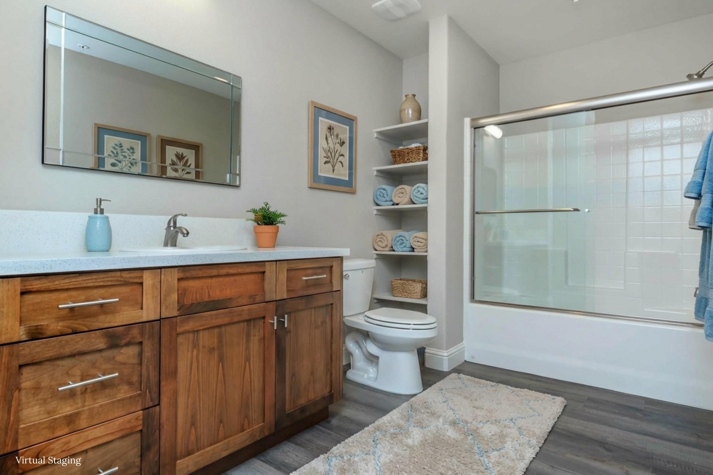 A well-lit modern bathroom featuring a wooden vanity with a sink, a large mirror above it, and a potted plant. There is a neatly organized shelf with rolled towels, a toilet, and a glass shower enclosure. The decor includes framed nature prints on the walls. Soft rug on the floor adds warmth to the space.