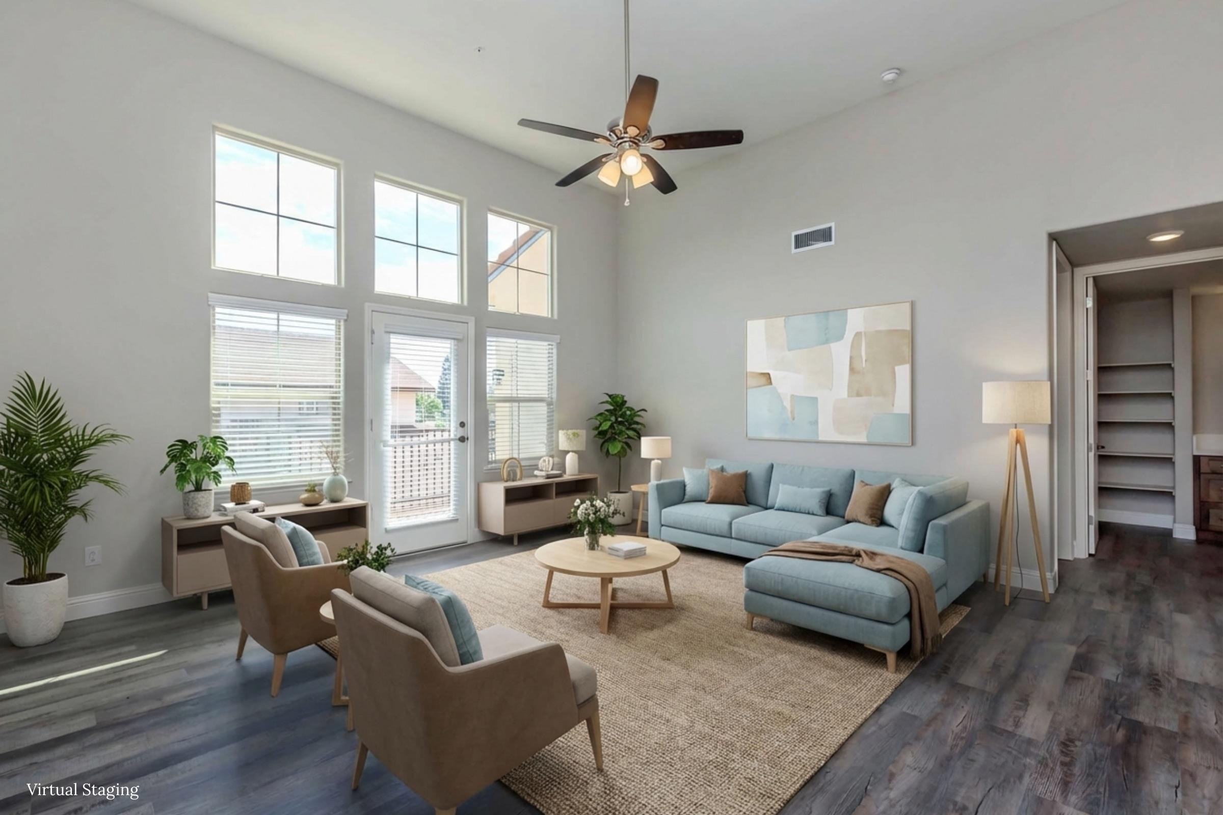 Modern living room featuring a light blue sofa, two beige armchairs, and a round coffee table. The space is well-lit with large windows and a ceiling fan. Decorative plants and minimalist artwork add a stylish touch. The floor is wooden, enhancing the open and airy feel of the room.