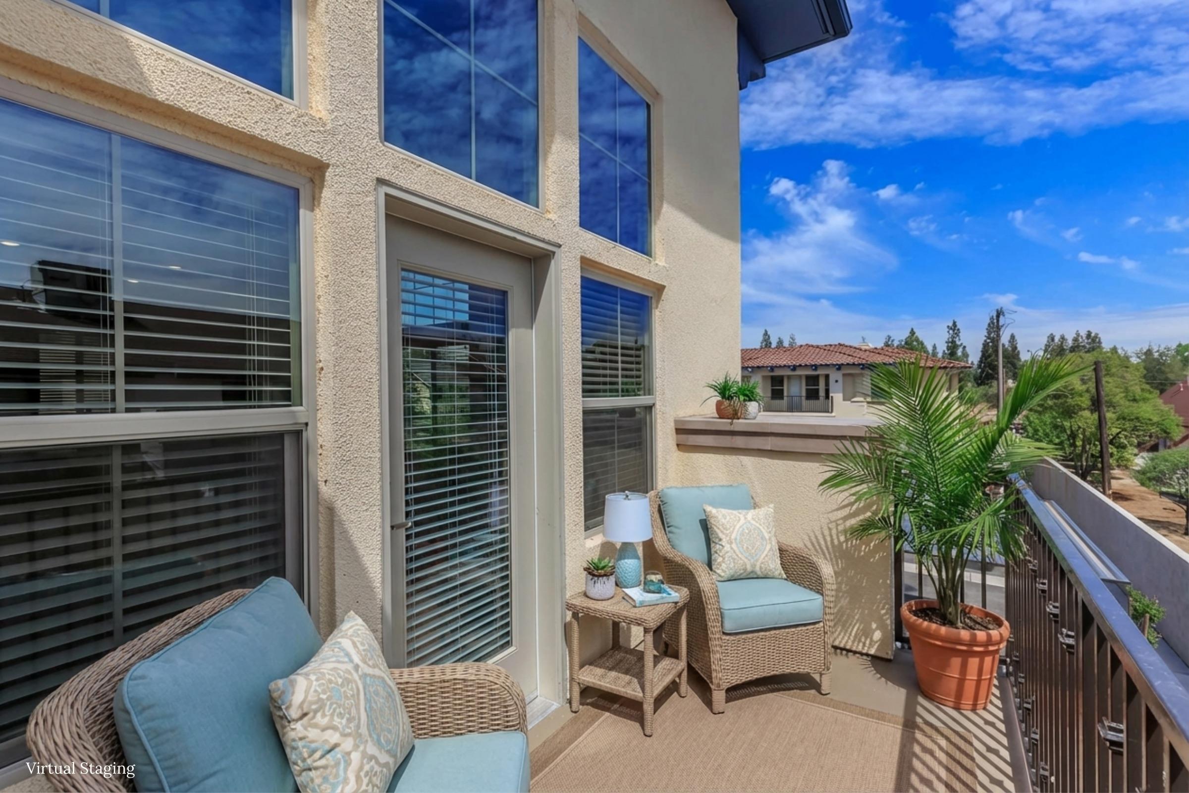 A sunlit balcony featuring two comfortable chairs with decorative pillows, a small table with a lamp, and a potted plant. Large windows offer a view of a scenic outdoor landscape with trees and blue sky. The space is inviting and ideal for relaxation.