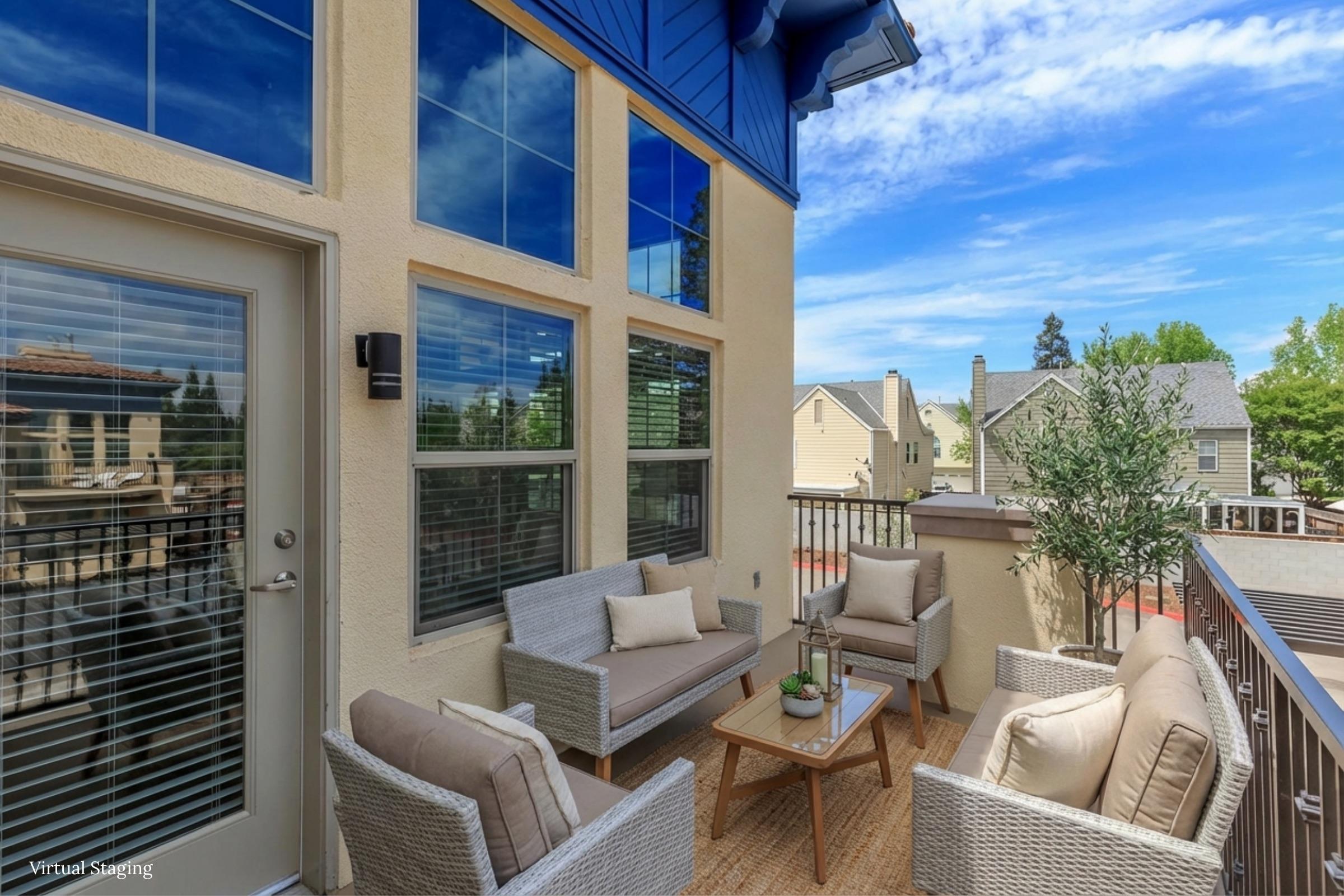 A cozy balcony with modern outdoor furniture, including a table and four cushioned chairs. Sunlight filters through large windows, illuminating the space. Potted plants and a scenic view of a neighborhood and blue sky enhance the inviting atmosphere.