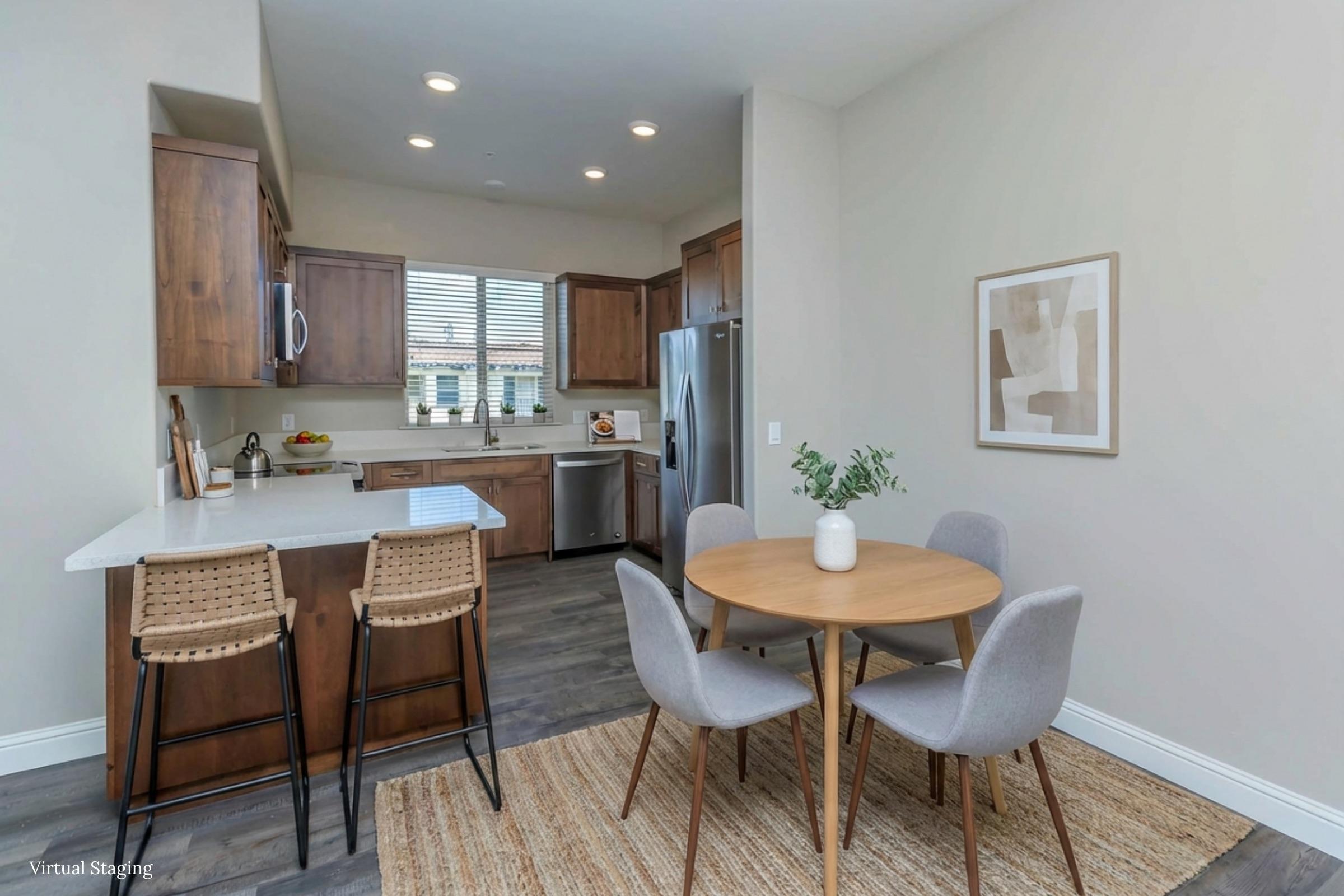 A modern kitchen featuring wooden cabinets, a white countertop, and stainless steel appliances. A small round dining table with four grey chairs is set in front of a large window, allowing natural light to brighten the area. A textured rug lies beneath the table, adding warmth to the space.