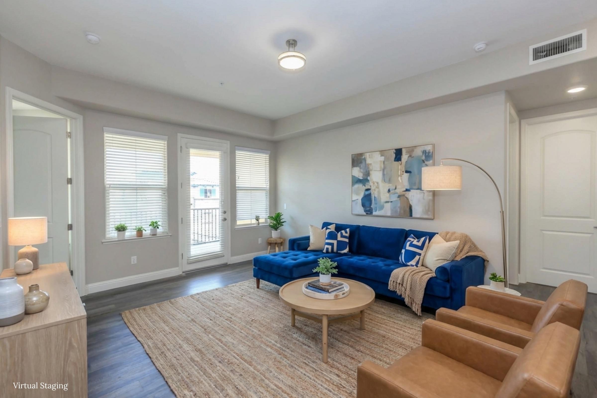 A cozy living room featuring a blue sofa with decorative pillows, a round coffee table, and two brown armchairs. Natural light streams in through large windows, and a stylish floor lamp adds warmth to the space. A textured area rug complements the modern decor.