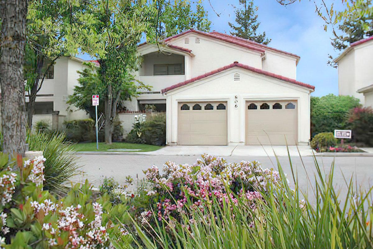 A suburban house with a light-colored exterior and a red-tiled roof, featuring a two-car garage. The front garden has various flowering plants and green grass, with trees in the background and a clear blue sky. A street sign is visible, indicating a residential area.