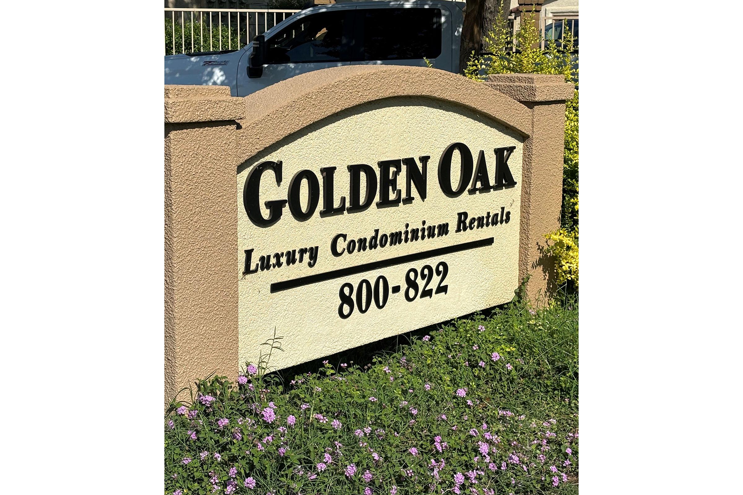 Sign for "Golden Oak Luxury Condominium Rentals" with a range of units from 800 to 822. The sign features large, bold lettering and is surrounded by greenery and flowering plants. The setting appears sunny and welcoming.