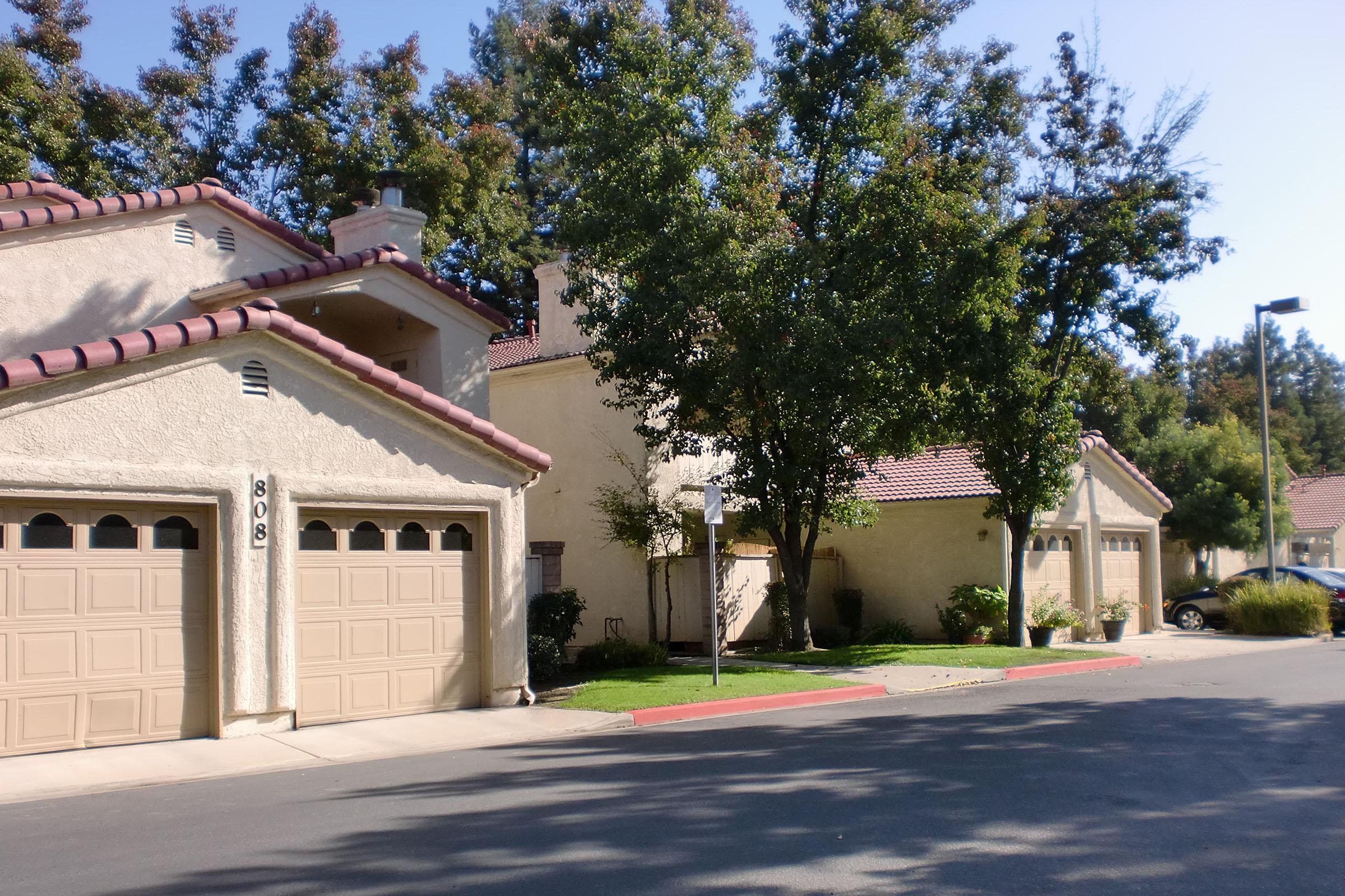 A quiet residential neighborhood featuring beige townhouse-style buildings with red tile roofs. Parking spaces are visible in front of the garages, and trees provide shade along the street. The scene is bright and sunny, with a clear blue sky overhead.