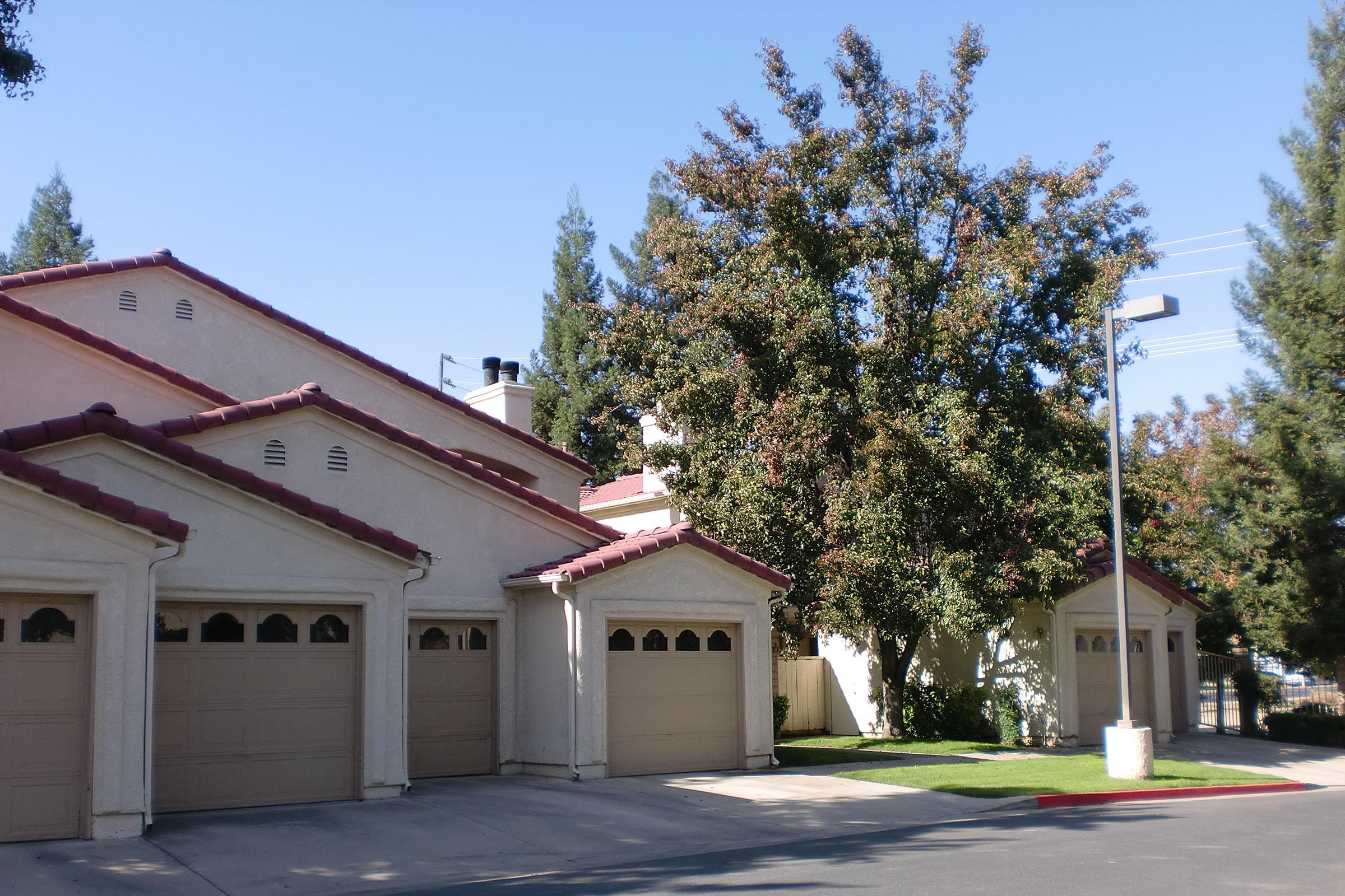 A row of tan residential garages with arched doors and maroon roofs, set against a backdrop of green trees and a clear blue sky. A small utility pole is visible on the right side of the image, providing street lighting. The grass is well-maintained, enhancing the suburban setting.