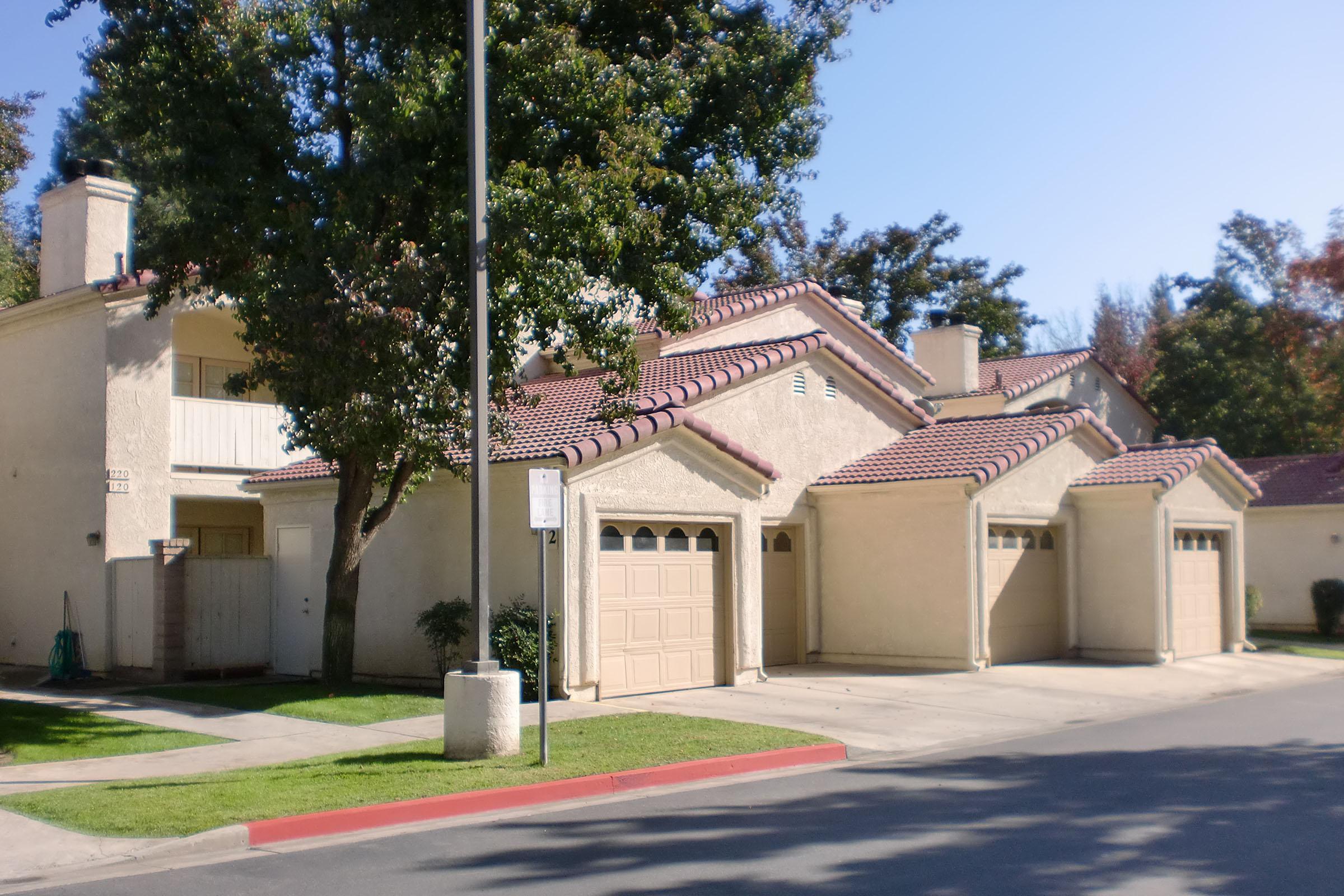 A residential scene featuring two-story homes with tan stucco exteriors and red tile roofs. The foreground includes a driveway with garage doors and a well-maintained lawn, while trees provide shade. The setting is bright and sunny, typical of a suburban neighborhood.