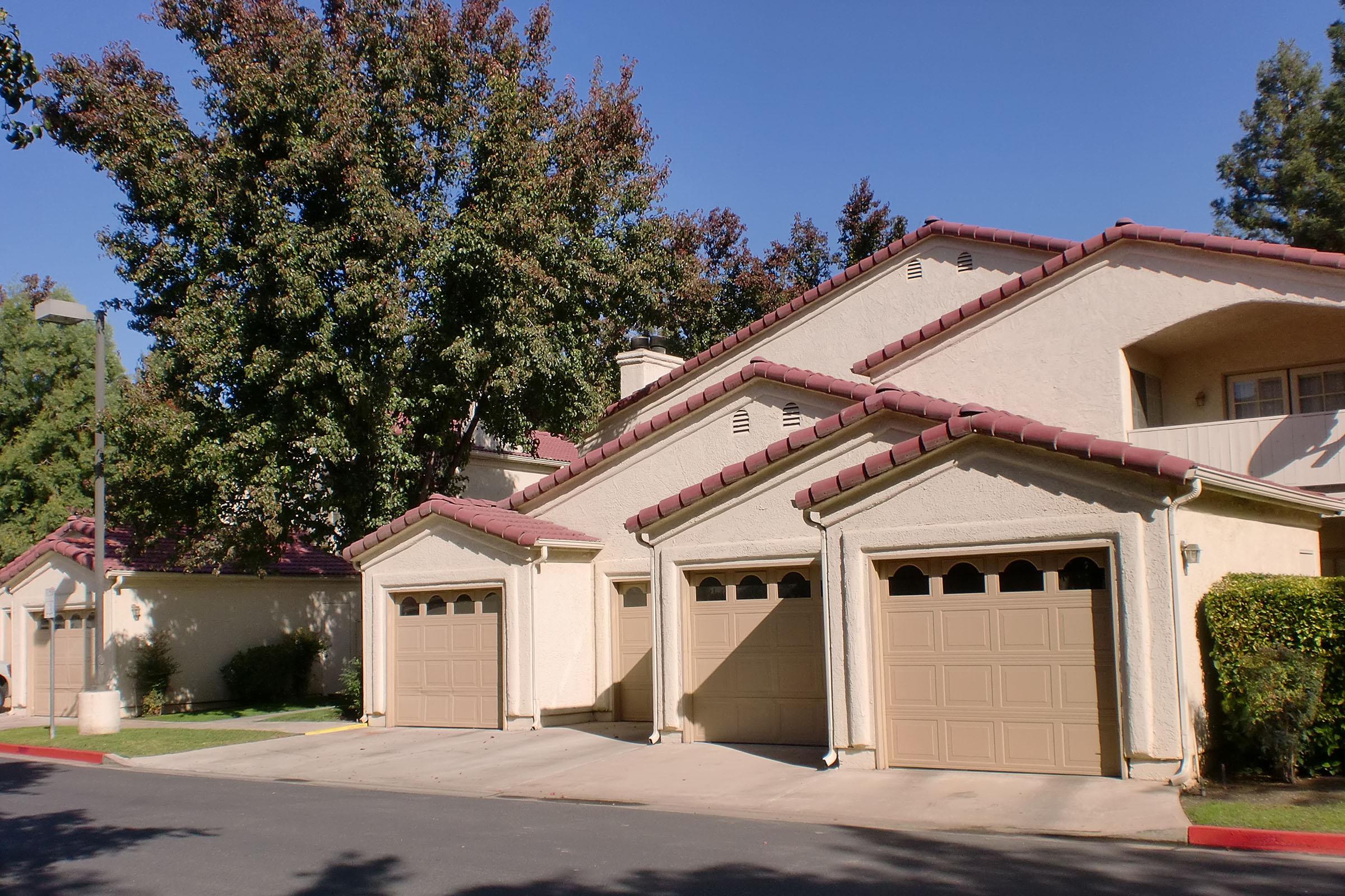 A residential building featuring a beige exterior with multiple garages, brown tiled roofs, and greenery. A large tree casts shade near the driveway, and a clear blue sky is visible in the background, indicating a sunny day.