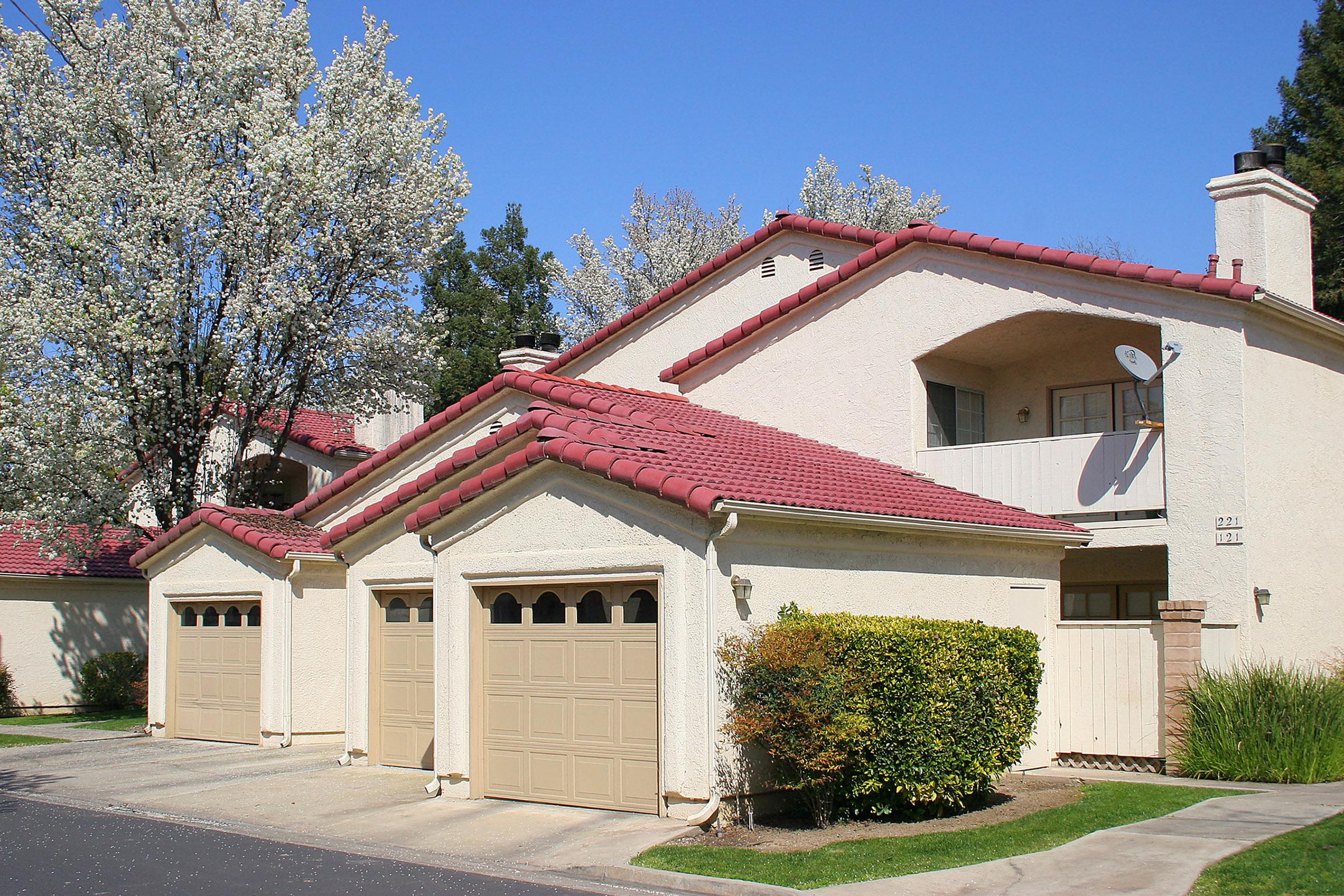 A suburban house with creamy-colored exterior walls and a red-tiled roof. It features two garage doors and a balcony with a satellite dish. Flanking the house are blooming trees, and the driveway leads up to the garages. Bright blue sky serves as the backdrop.