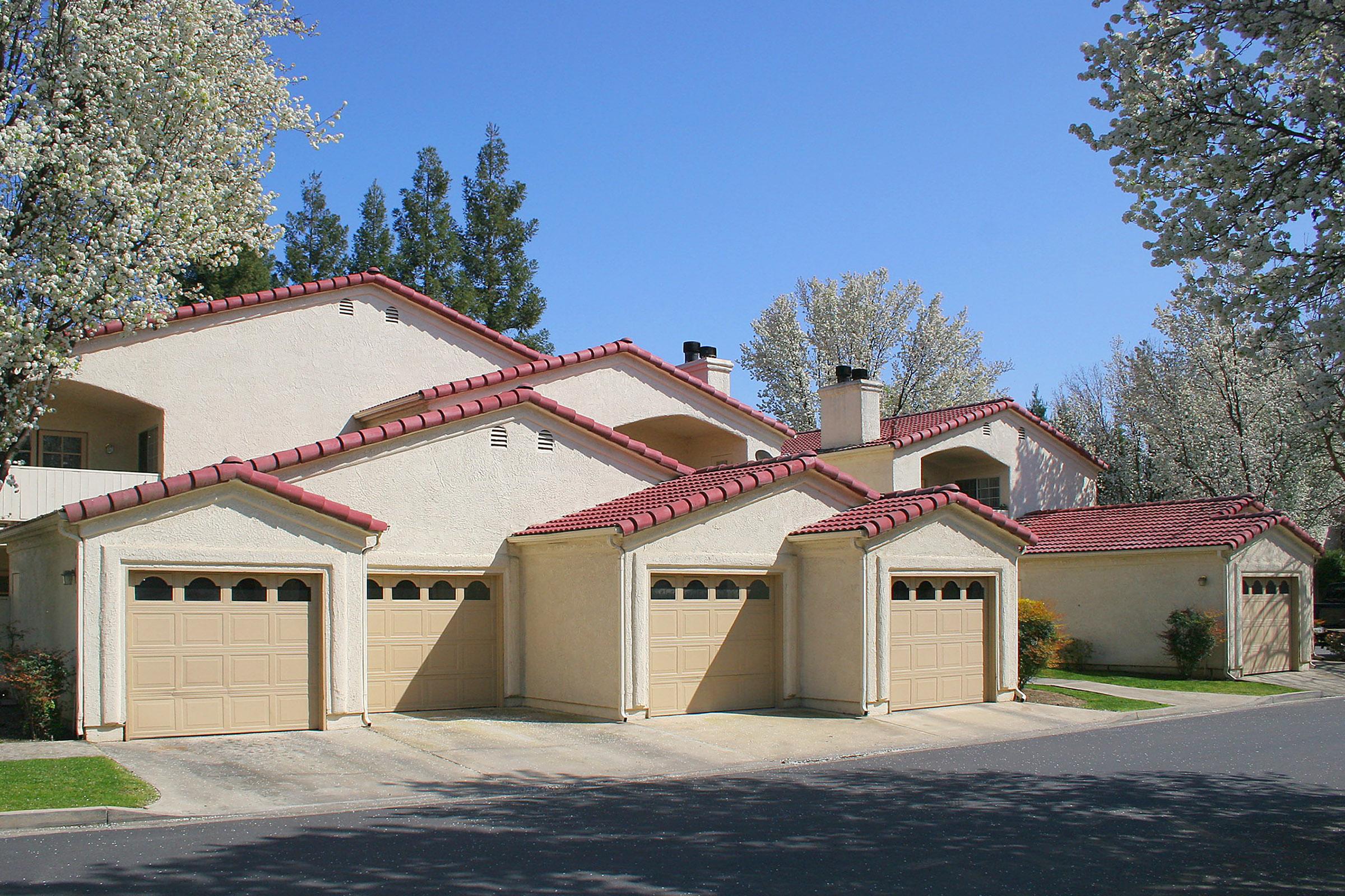 A residential neighborhood with several light-colored houses featuring red tile roofs and attached garages. Lush green trees and flowering plants line the street under a clear blue sky, creating a pleasant spring atmosphere.