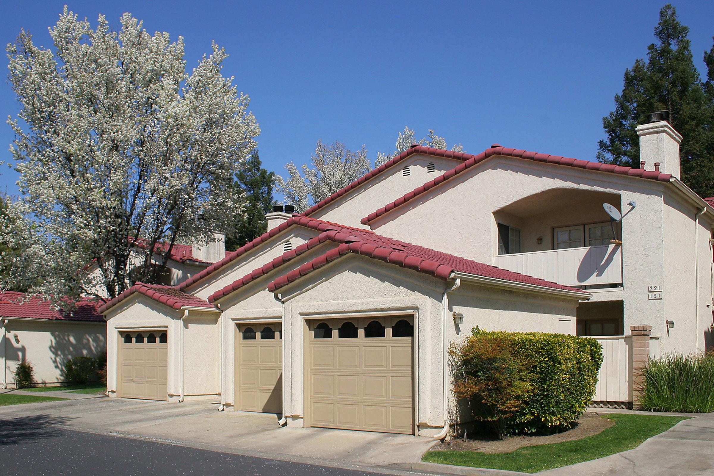 A picturesque residential scene featuring a two-story apartment building with a terra-cotta tiled roof. The building has multiple garage doors and is surrounded by well-maintained landscaping, including a flowering tree and lush greenery under a clear blue sky.