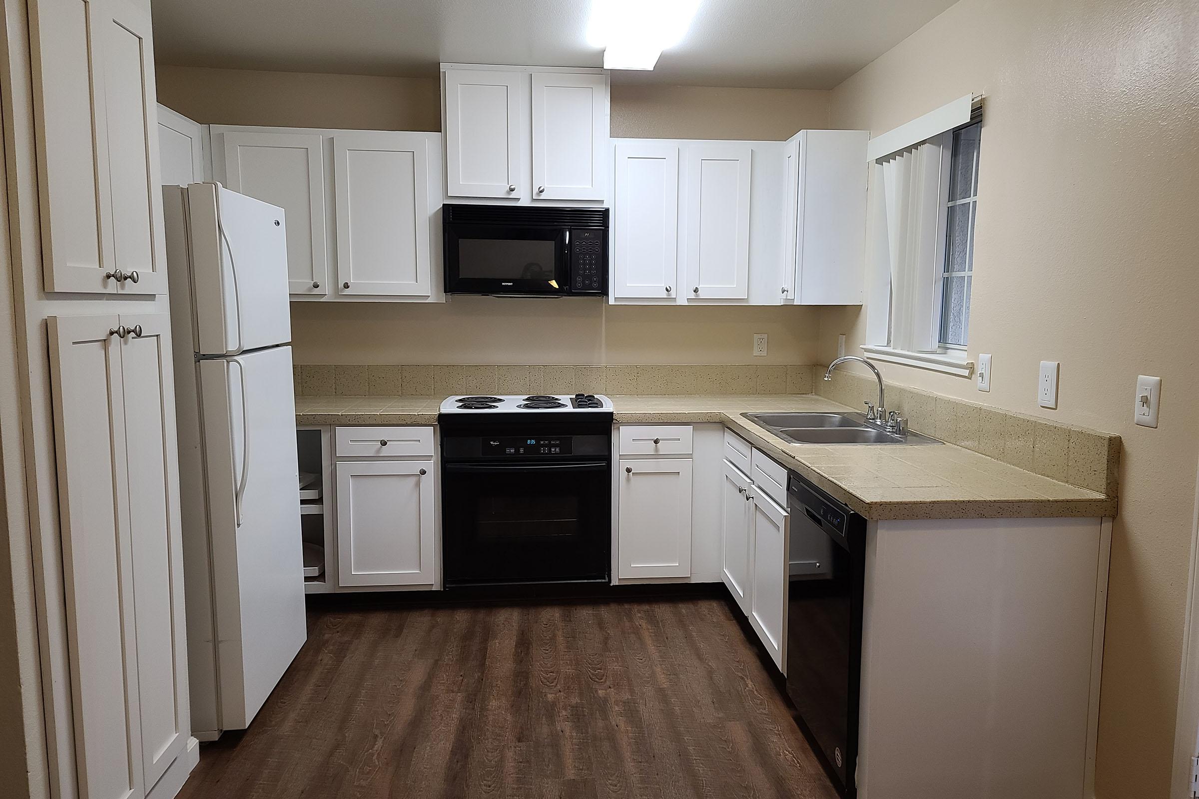 A modern kitchen featuring white cabinets, a black stove, and a microwave. It includes a refrigerator, a dishwasher, and a double sink with a window above it. The countertop is beige, and the floor is a dark wood laminate. Bright overhead lighting illuminates the space.