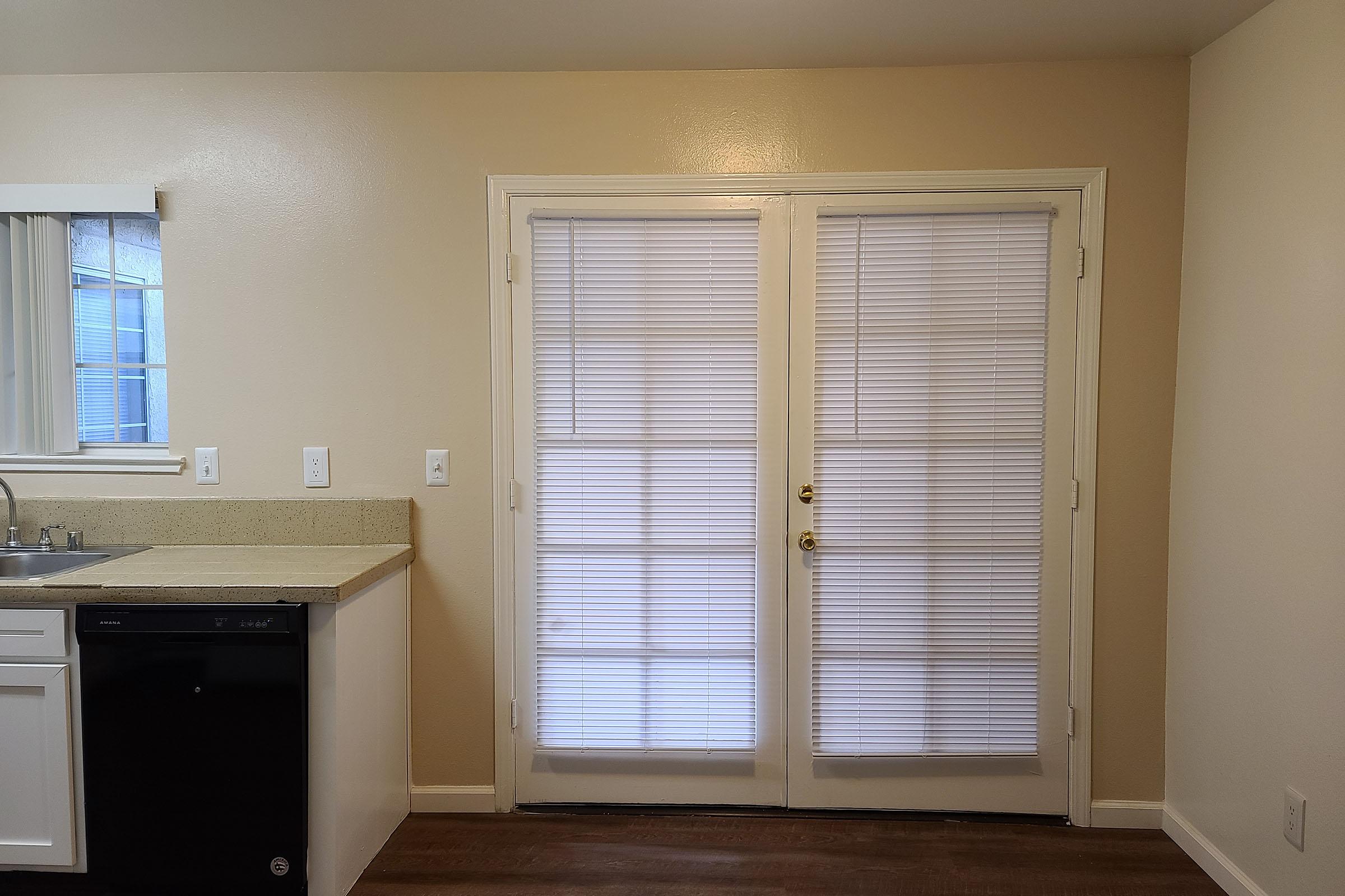 A kitchen space featuring a countertop with a sink and a black dishwasher. To the right, double sliding glass doors with white blinds open to an outside area, while the walls are painted a light beige color. The flooring is dark wood laminate.