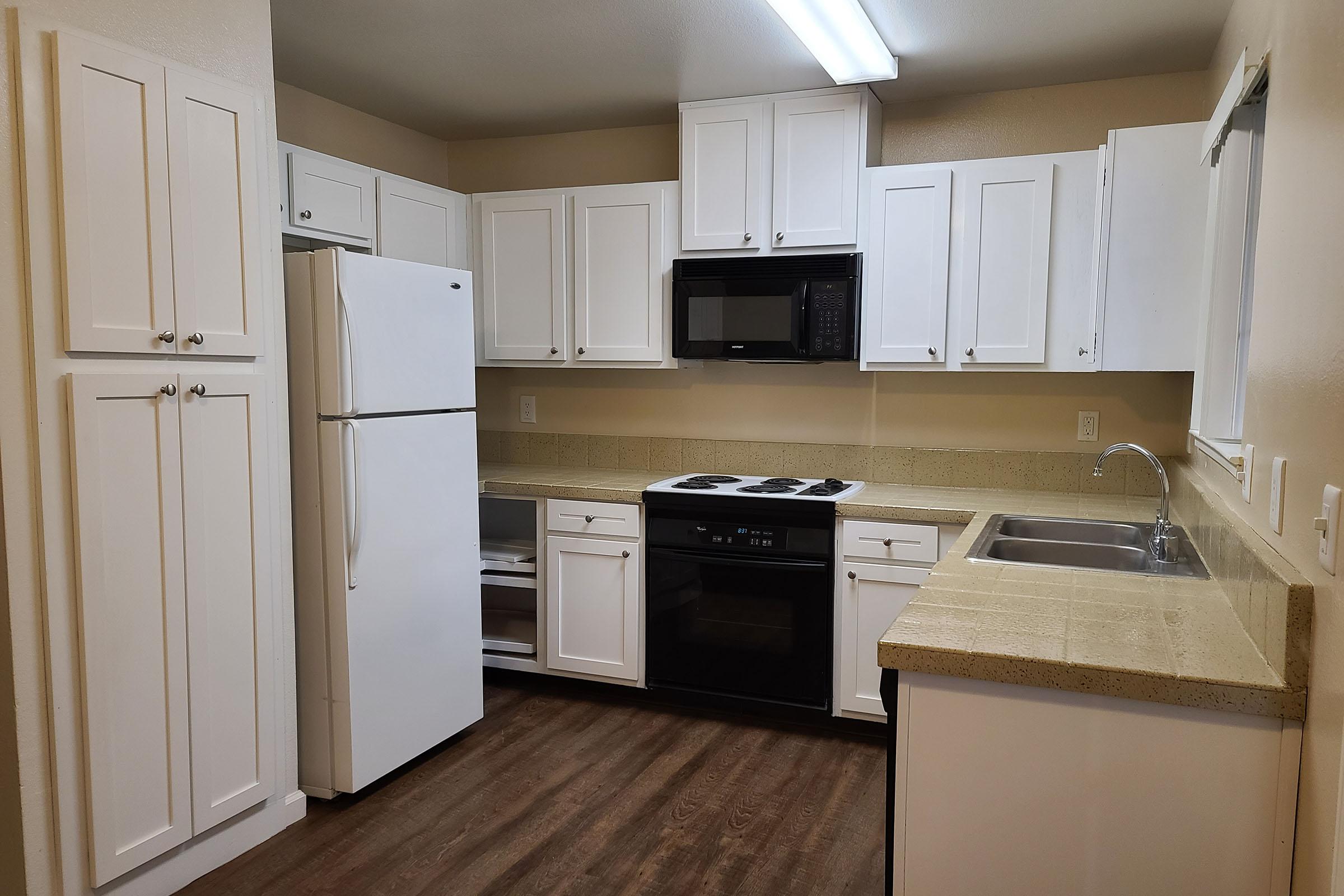 A modern kitchen featuring white cabinets, a large refrigerator, a microwave, and a black stove. The countertop is beige, with a sink situated next to it. The kitchen has a neutral color scheme and offers ample storage with multiple cabinets. Bright overhead lighting enhances the inviting atmosphere.