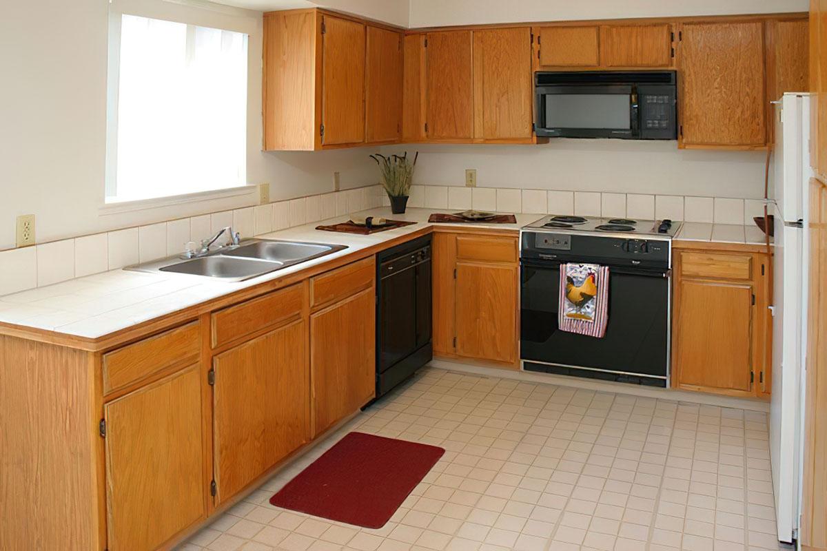 A modern kitchen featuring light wooden cabinets, a double sink, and a black microwave above a gas stove. The kitchen includes a dishwasher, a window allowing natural light, a tiled countertop, and a red mat on the floor. A decorative towel hangs on the stove.