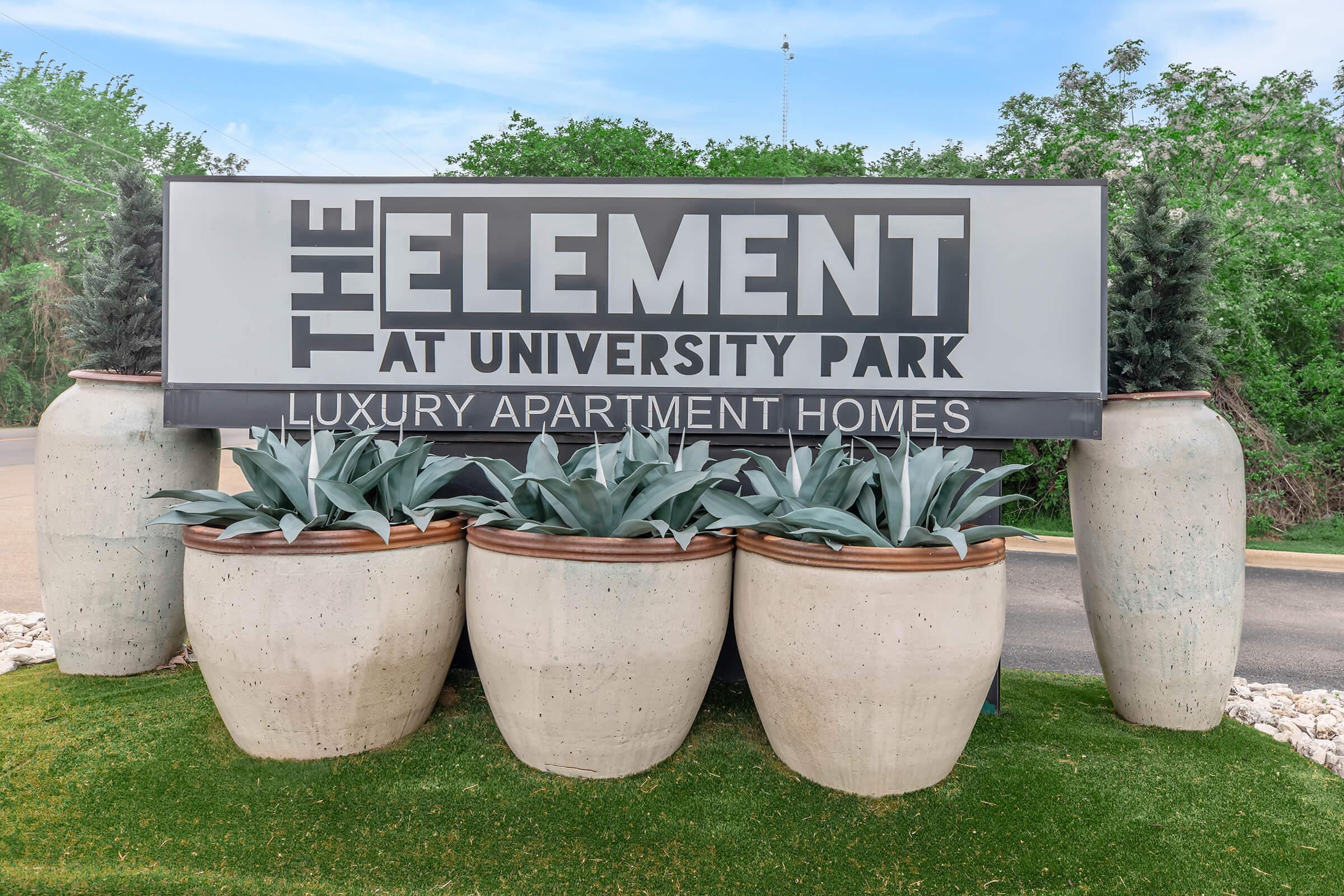 Sign for "The Element at University Park" featuring the text "Luxury Apartment Homes." The sign is surrounded by large decorative planters with leafy plants, placed on a green lawn, set against a backdrop of trees and blue sky.