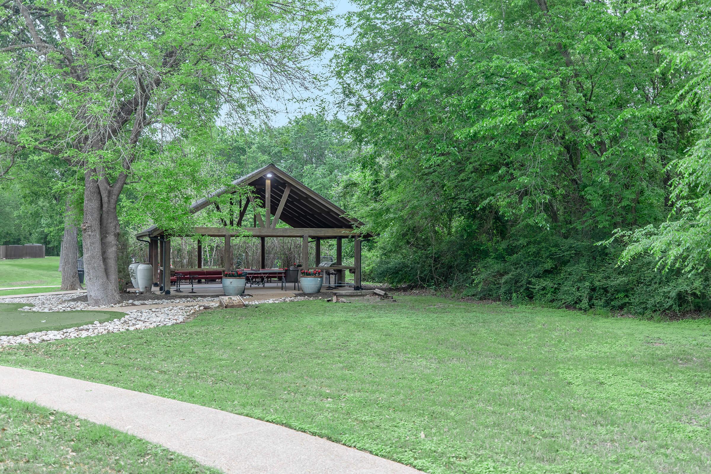 A wooden pavilion with a thatched roof is nestled among lush greenery, surrounded by a well-maintained lawn. Stone pathways lead to the pavilion, which features a shaded seating area. Trees provide a natural backdrop, creating a serene and inviting outdoor space.